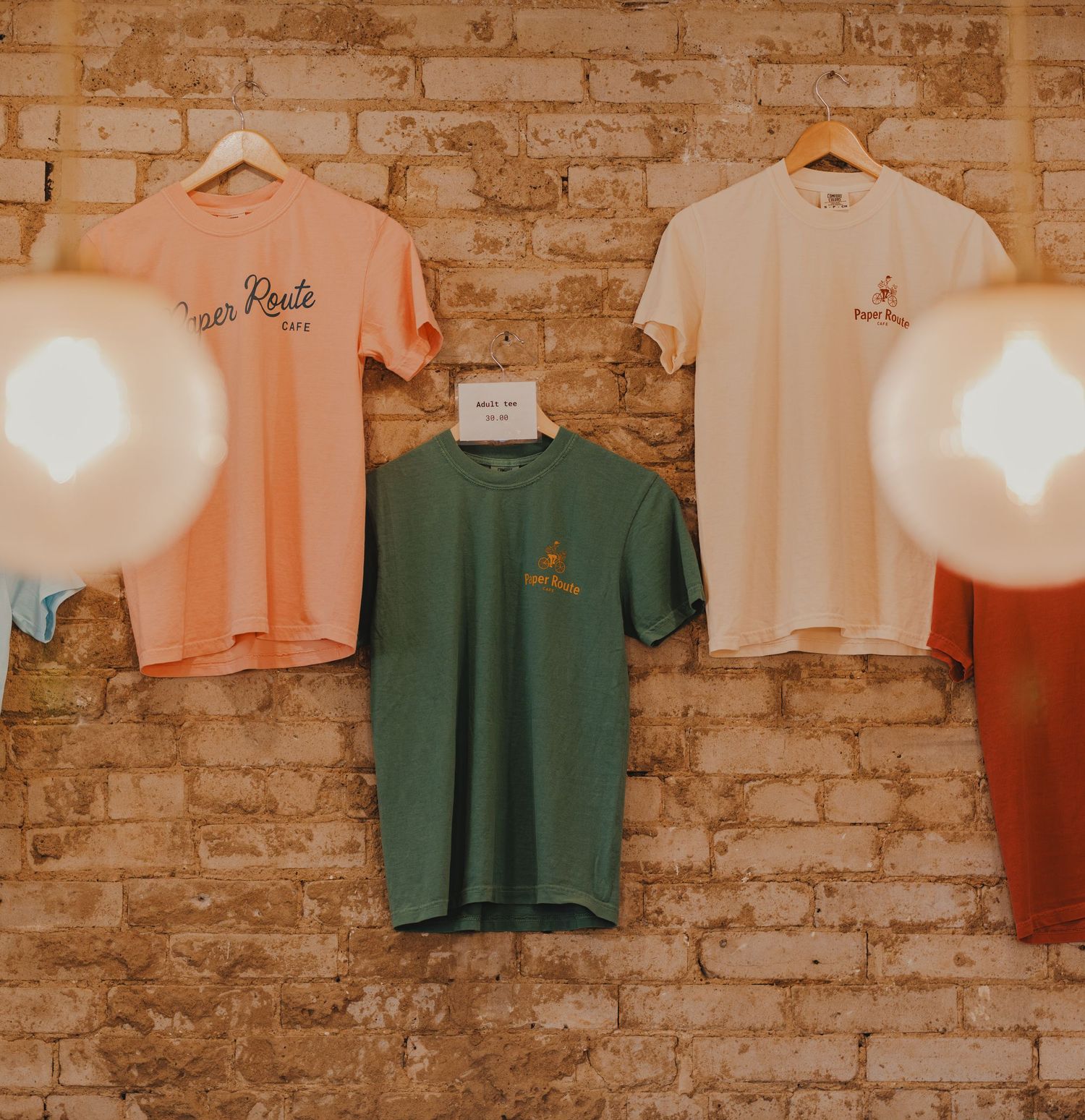 Three t-shirts in peach, green, and cream hanging on a rustic brick wall with warm globe lights.