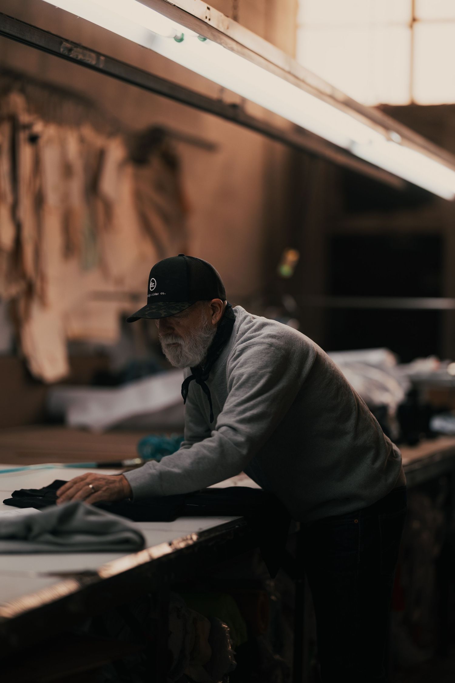 Elderly craftsman in black cap working with fabric at a workbench in a dimly lit textile workshop.