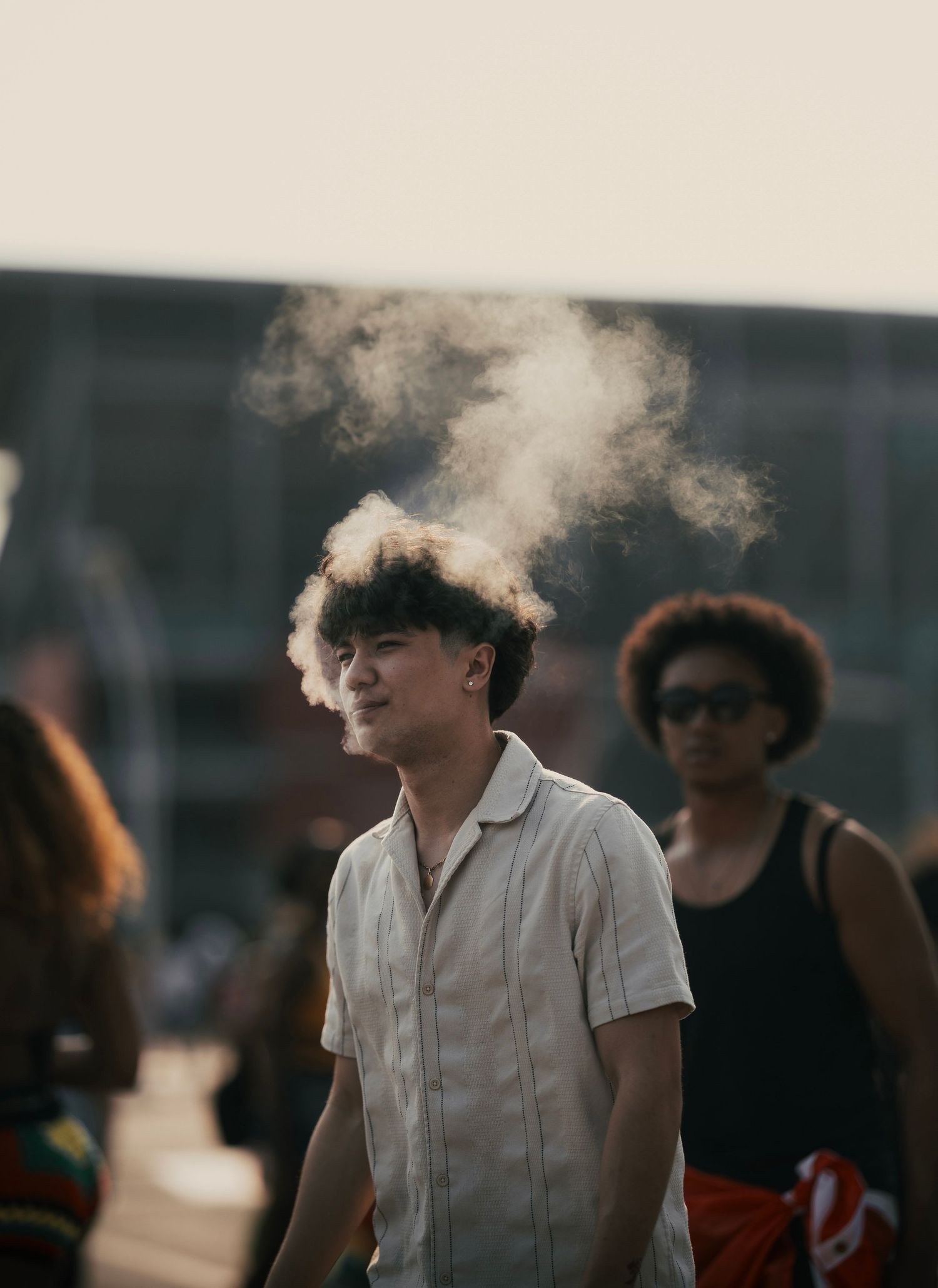 Young man exhaling vape smoke outdoors at a public event, with blurred people in the background.