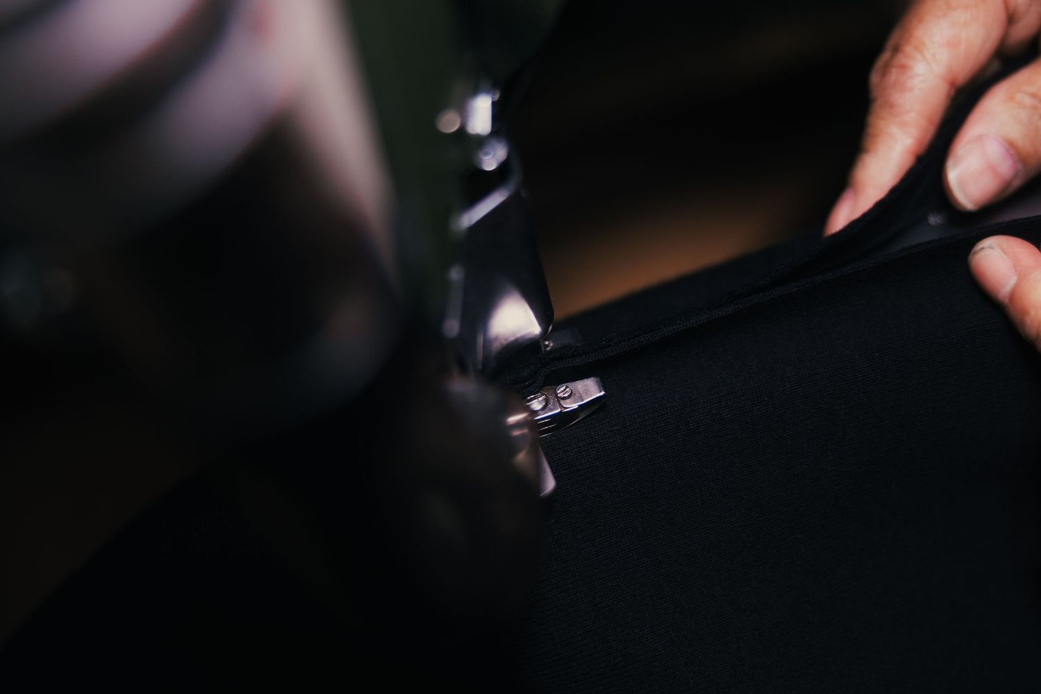 Close-up of hands using a zipper pull tool on a black fabric zipper in a dimly lit workspace.