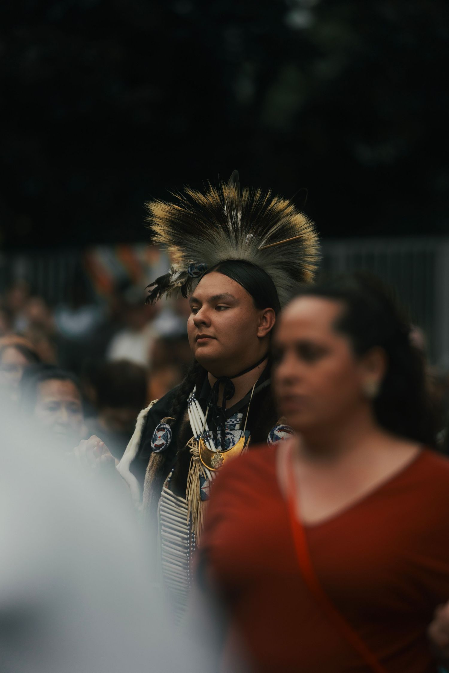 Native American man in traditional feathered headdress stands among crowd at outdoor cultural event.
