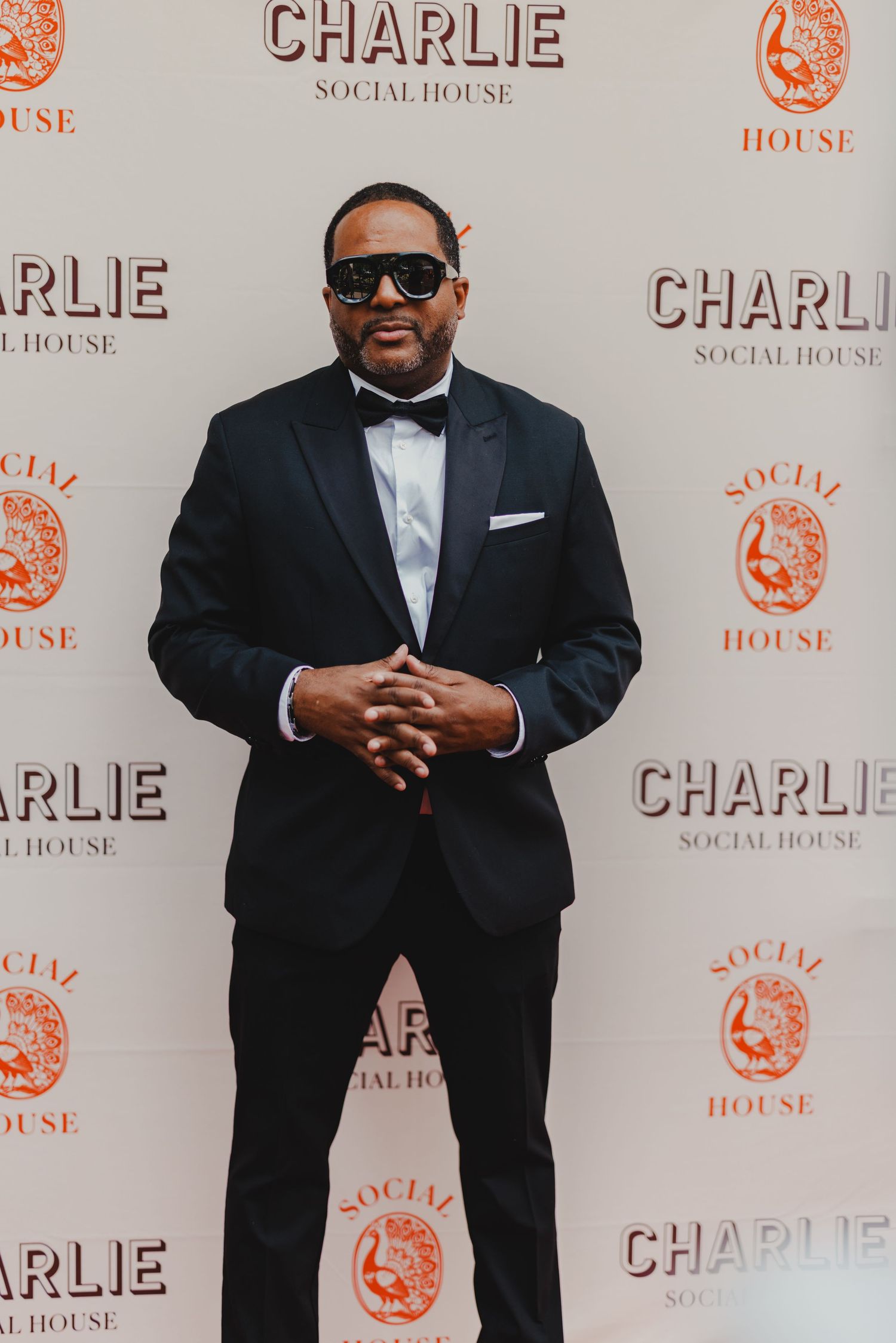 Man in black tuxedo with bow tie and sunglasses posing at Charlie Social House event on red carpet.