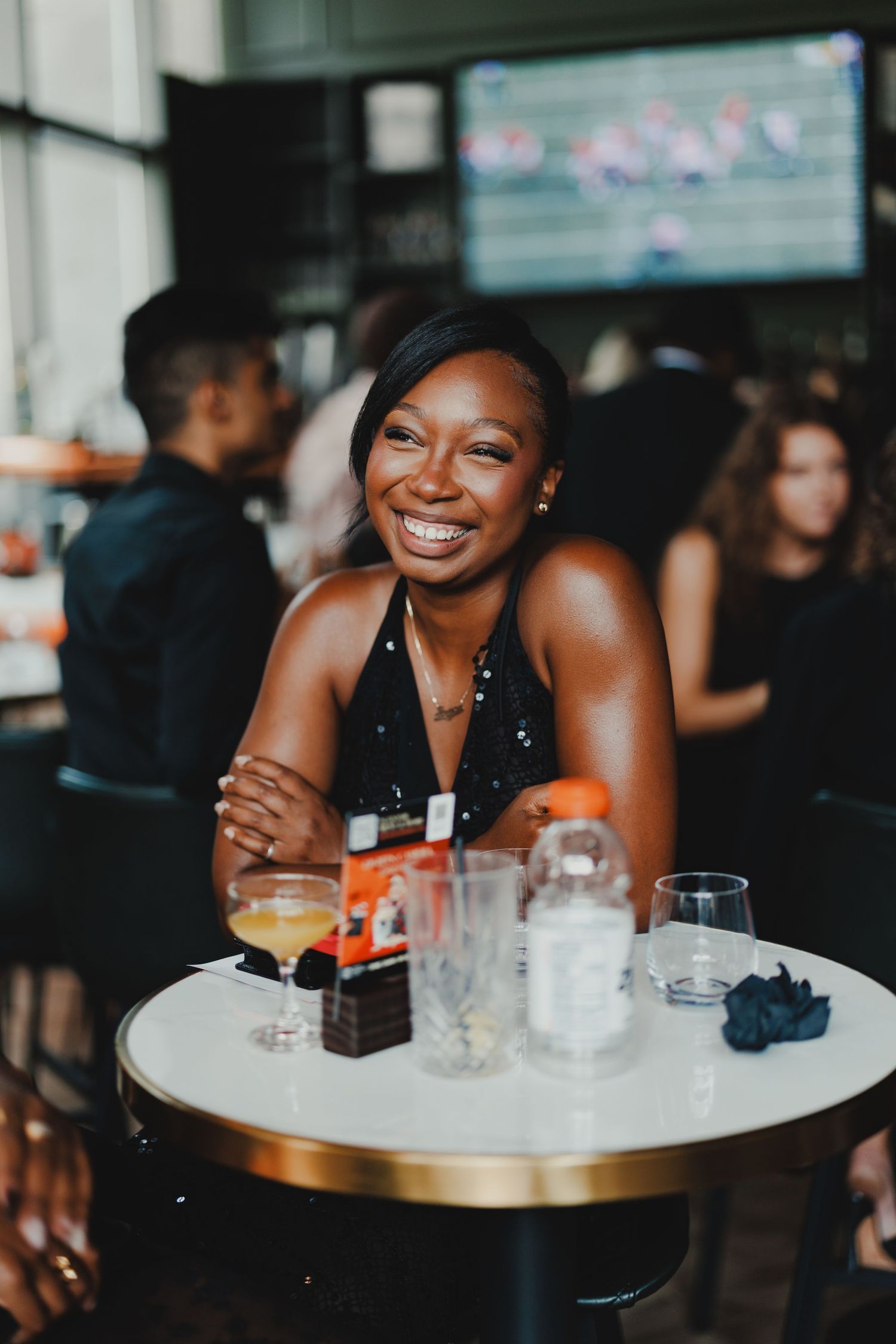 A smiling young woman in a black dress sits at a white bar table with cocktails, enjoying a social event.