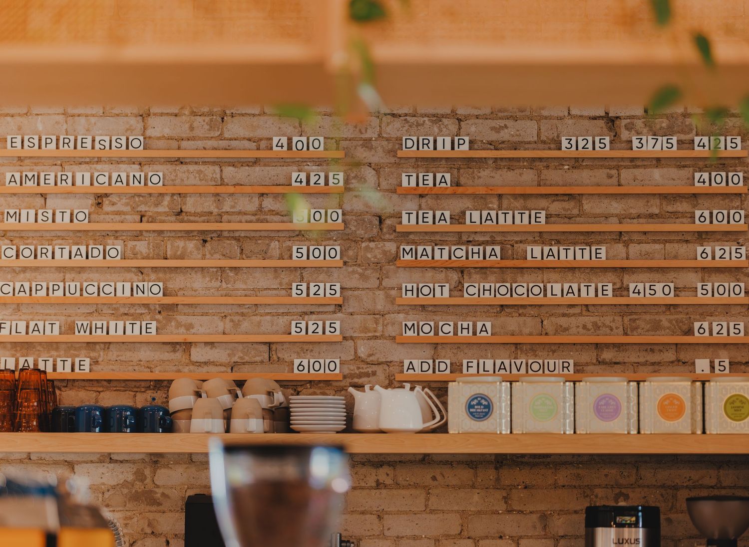 A cafe menu board on a brick wall listing coffee drinks and prices with colorful tile accents.