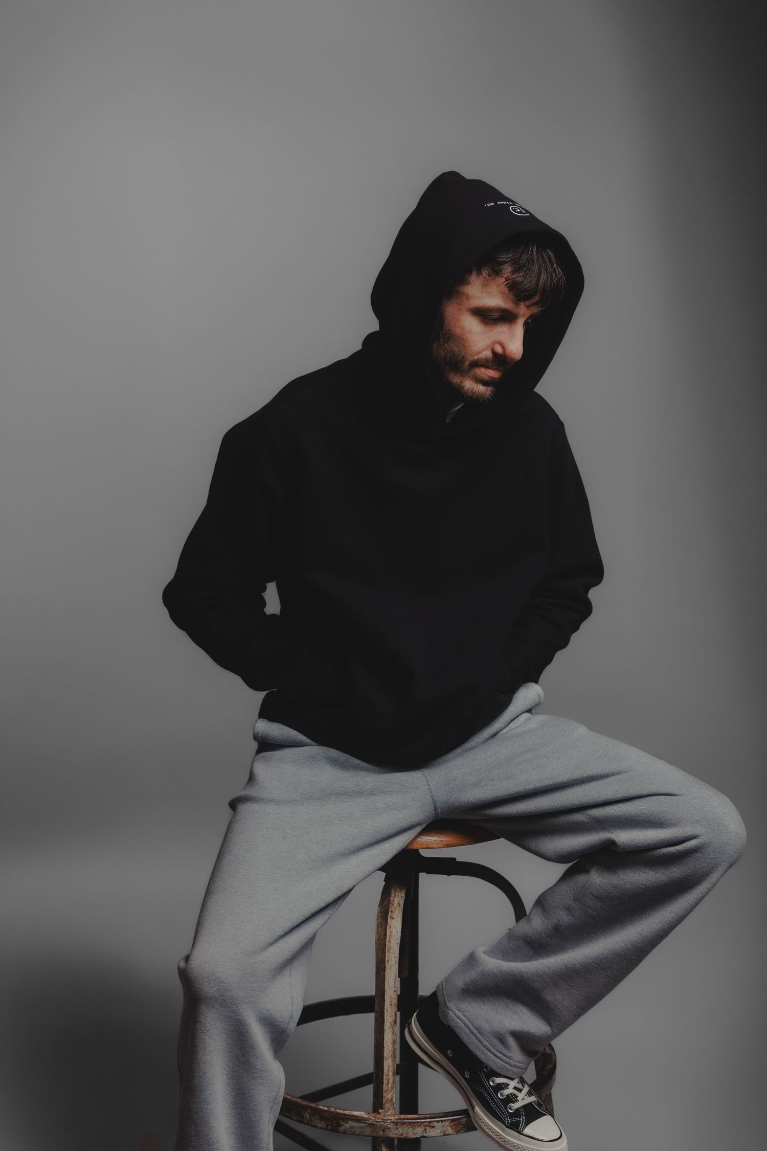 Bearded man in black hoodie and grey sweatpants sitting on stool in studio setting.