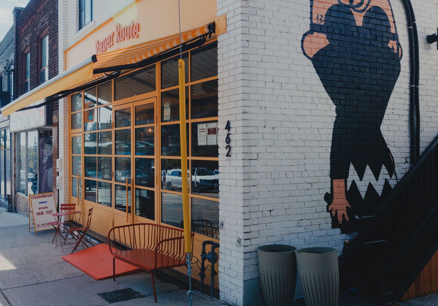 Exterior of a restaurant with yellow awning, red chairs, and a large street mural on a white brick corner building.
