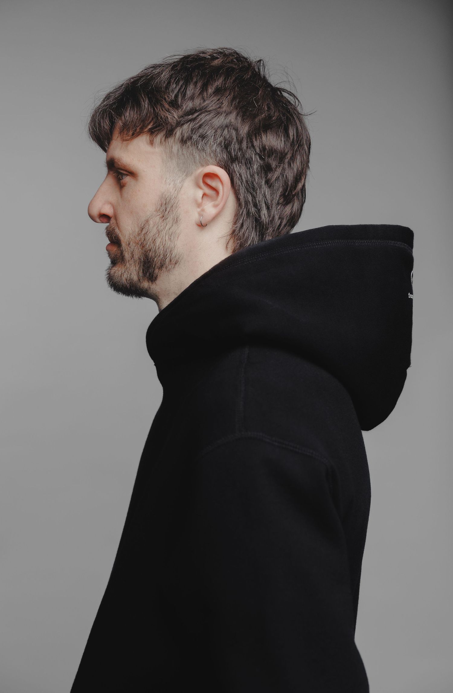 Man in profile view wearing a black hoodie with short dark hair and beard against gray background.