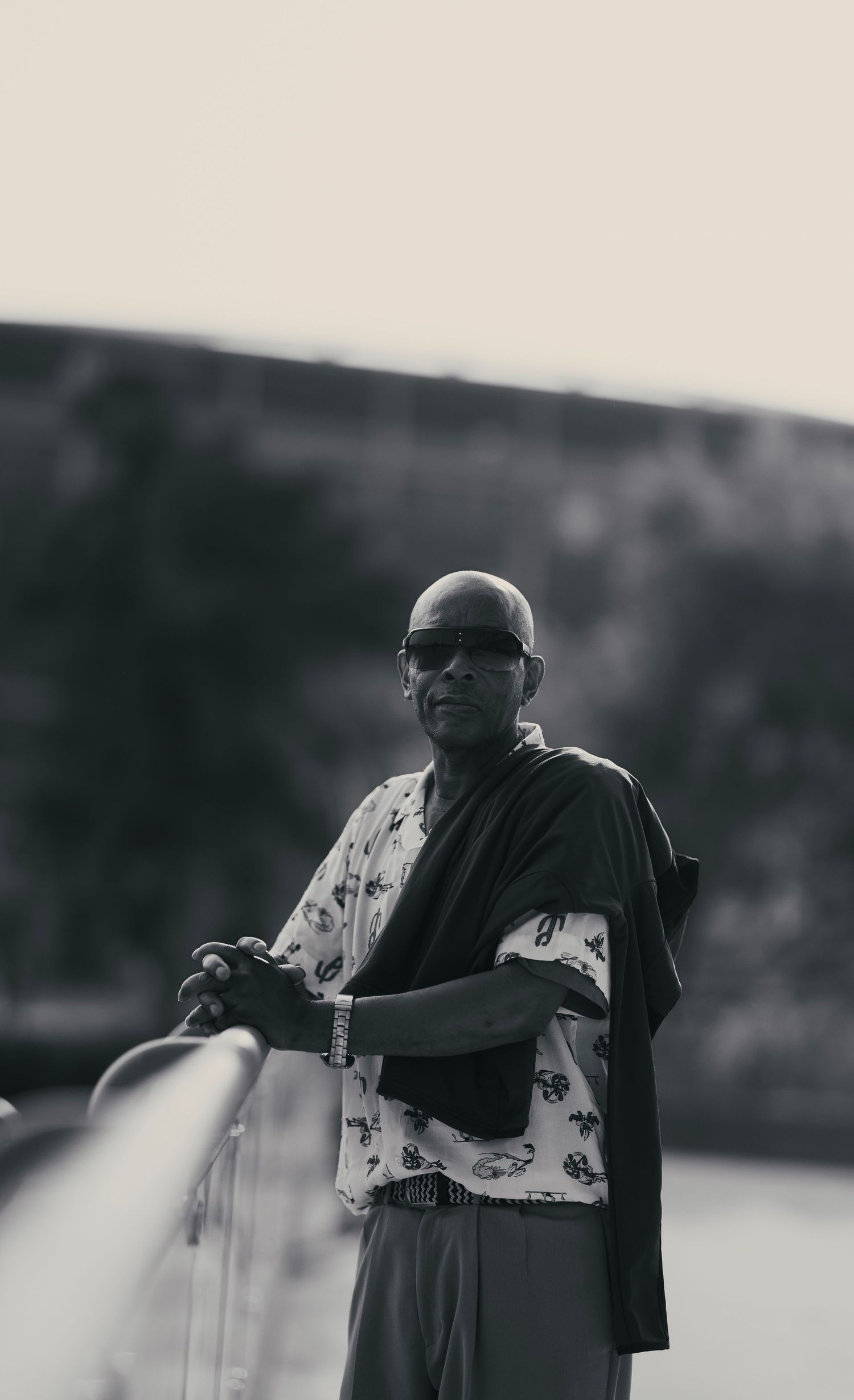 Bald man wearing sunglasses and a patterned shirt with a dark shawl, leaning on a railing in black and white photo.