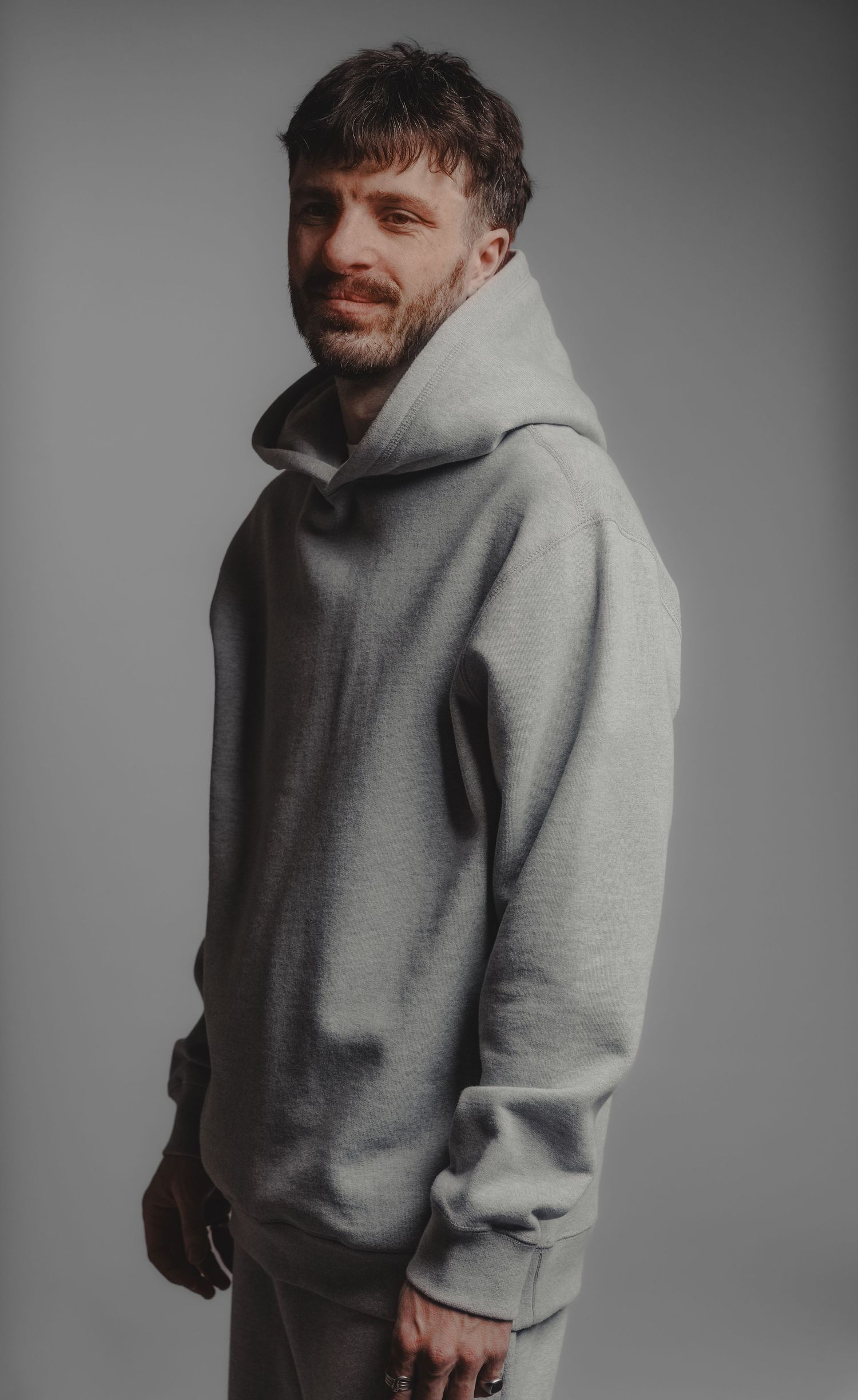 Bearded man smiling in a grey oversized hoodie and sweatpants, side profile studio shot.
