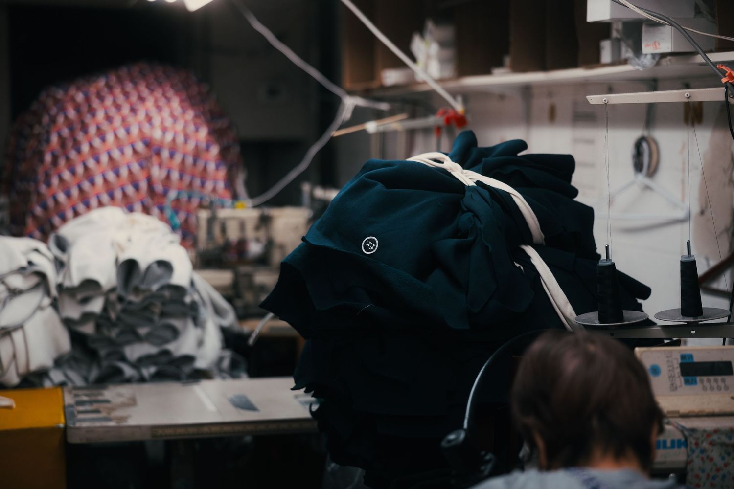 A busy garment workshop with dark fabric piled on a sewing machine table, colorful textile rolls visible in the background.