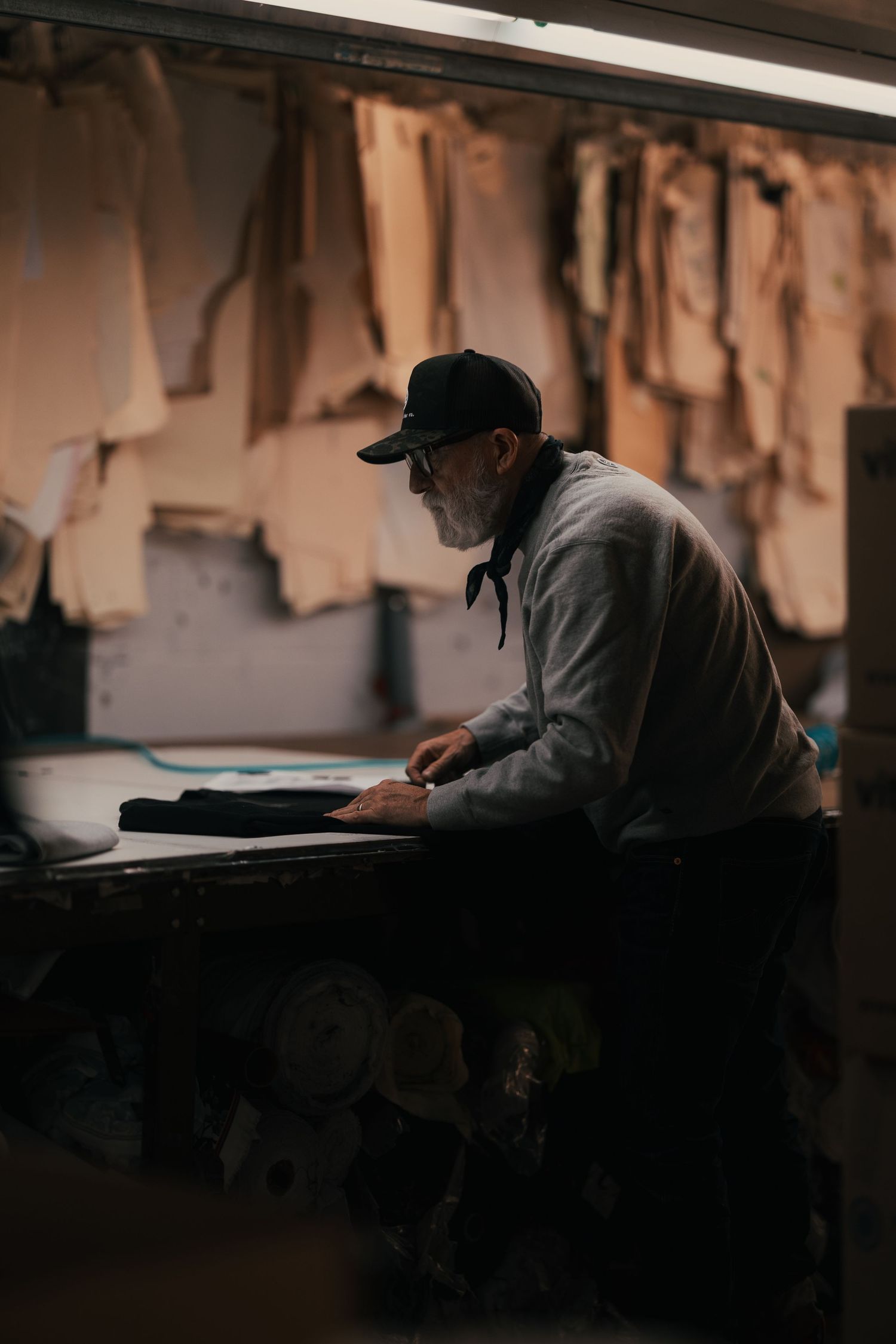 Elderly craftsman working at a leather workshop bench surrounded by hanging leather patterns in a dimly lit studio.