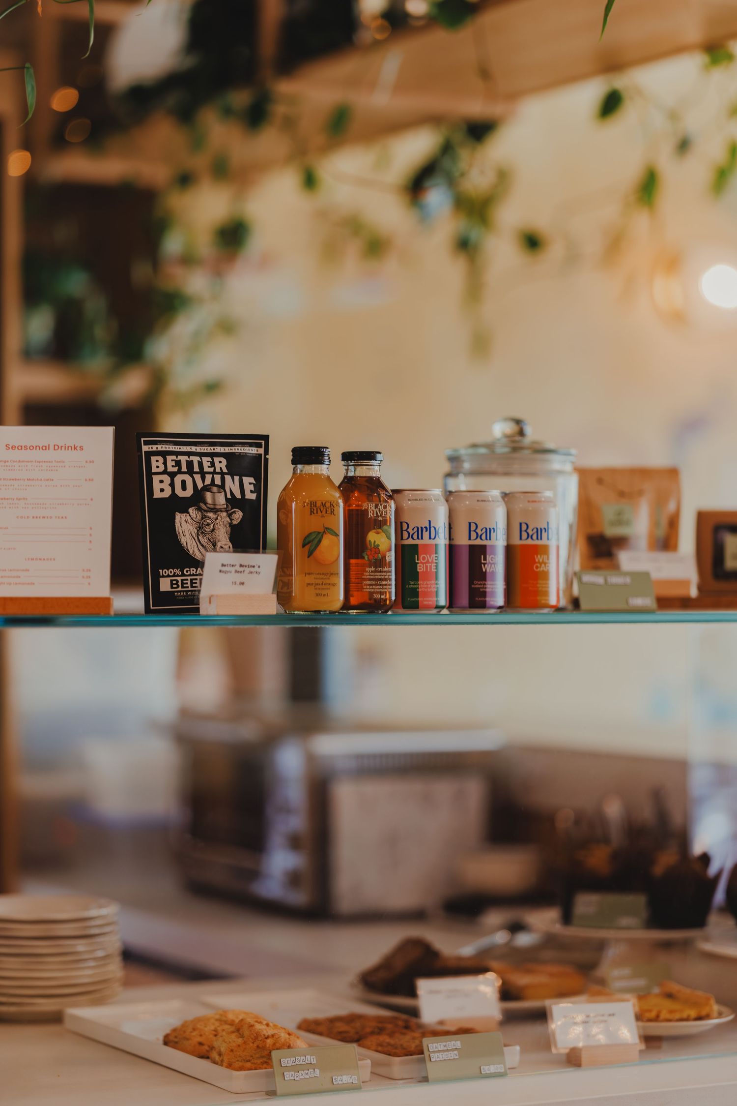 Coffee shop display shelf with Better Bovine products, bottled drinks, Barb cans, and freshly baked pastries.