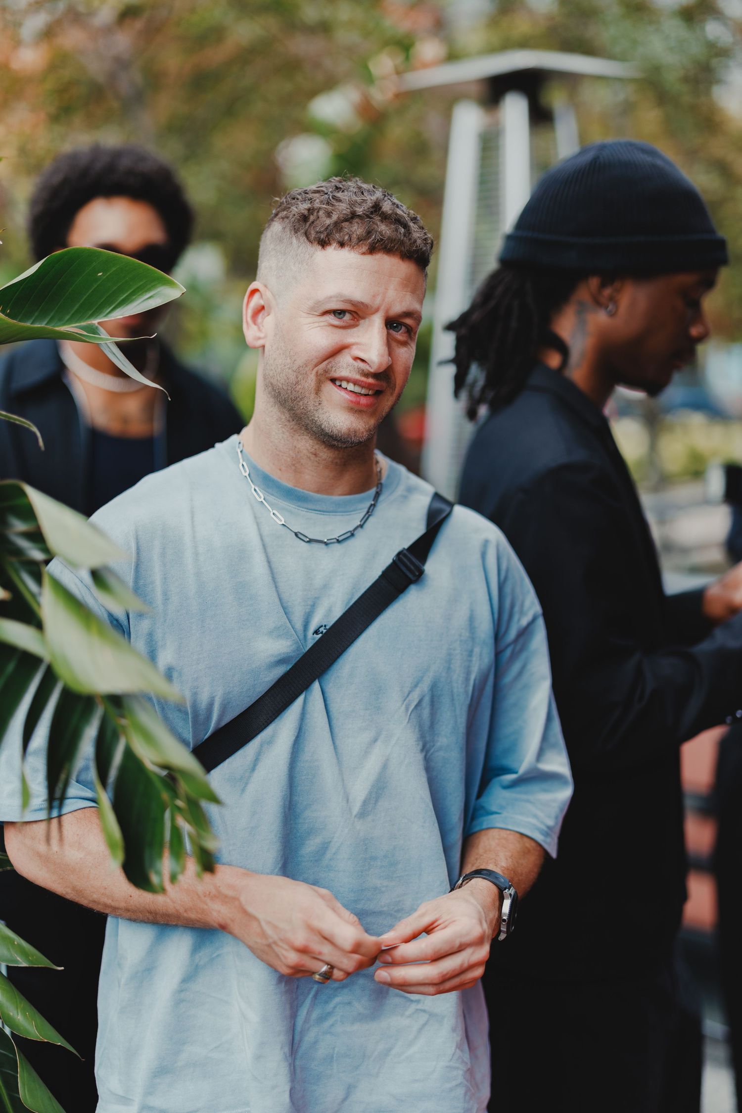 Smiling man in blue oversized t-shirt with crossbody bag stands outdoors among a diverse group of people.