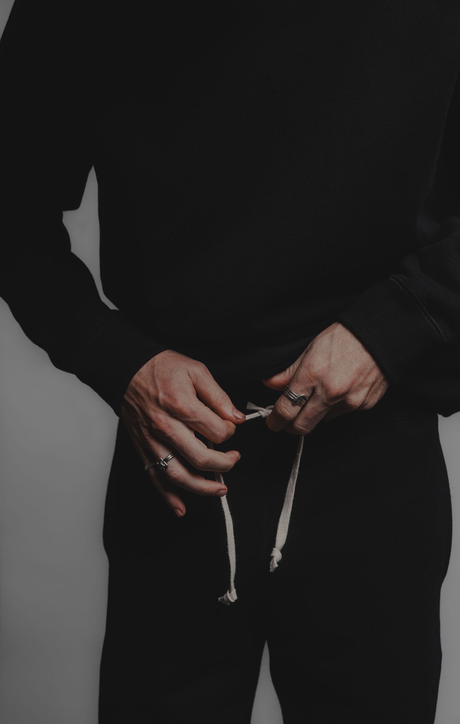 Man in black sweatshirt adjusting drawstring cord with ringed fingers against grey background.