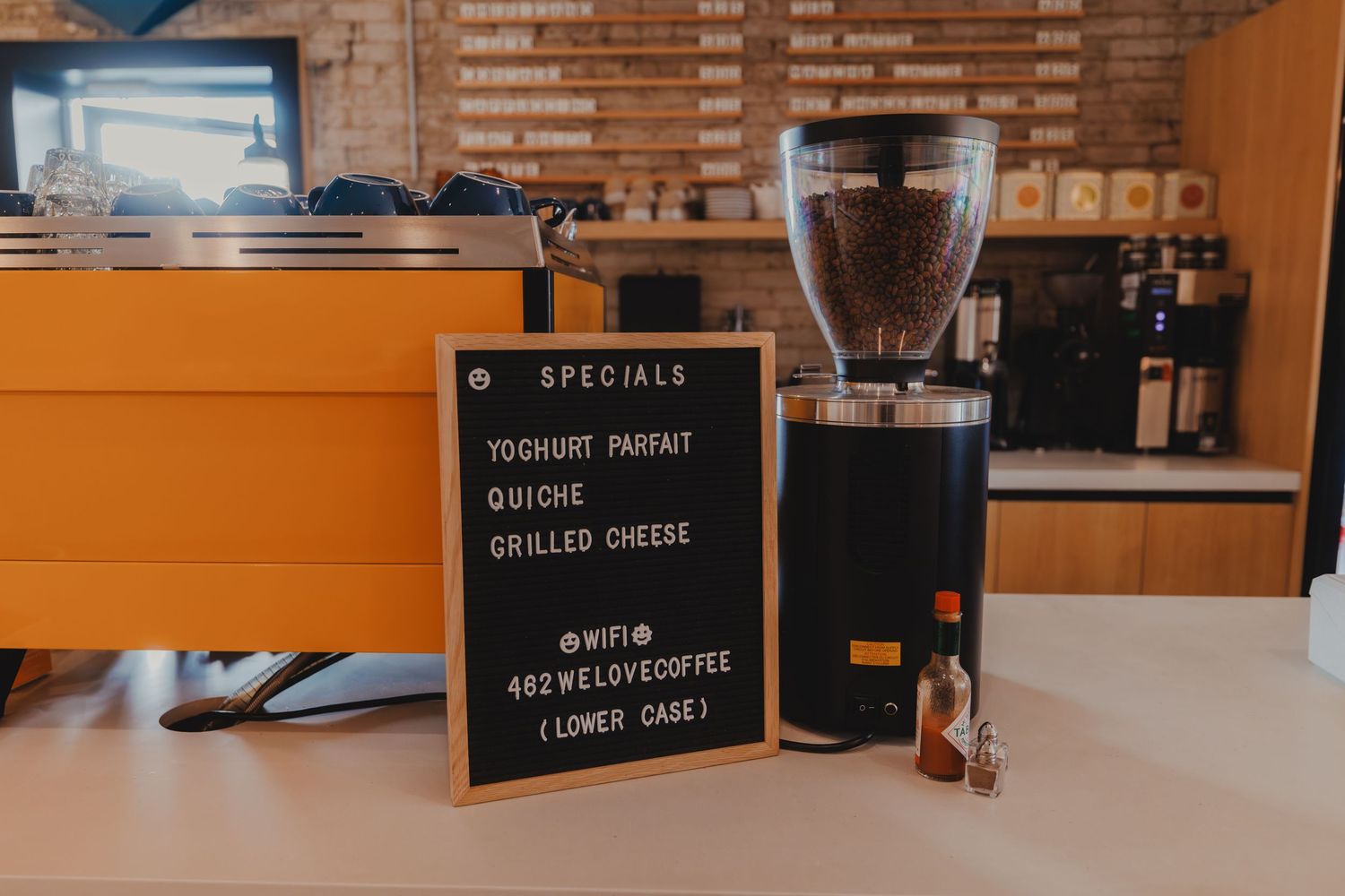 Coffee shop specials board listing yogurt parfait, quiche, and grilled cheese beside a black coffee bean grinder.