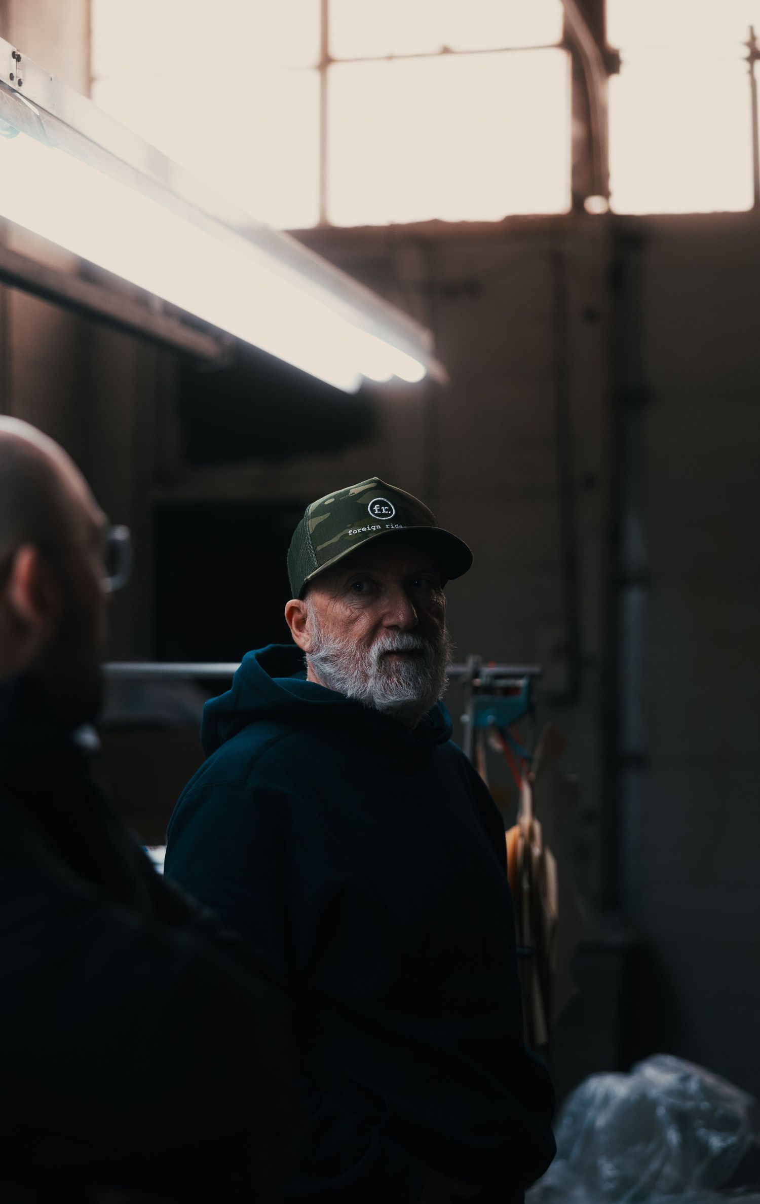 Elderly bearded man wearing a green cap standing in an industrial workspace with dramatic backlit window.