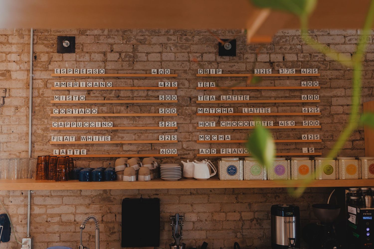 Rustic brick wall coffee shop menu board above wooden shelf with mugs and supplies in a cozy cafe interior.