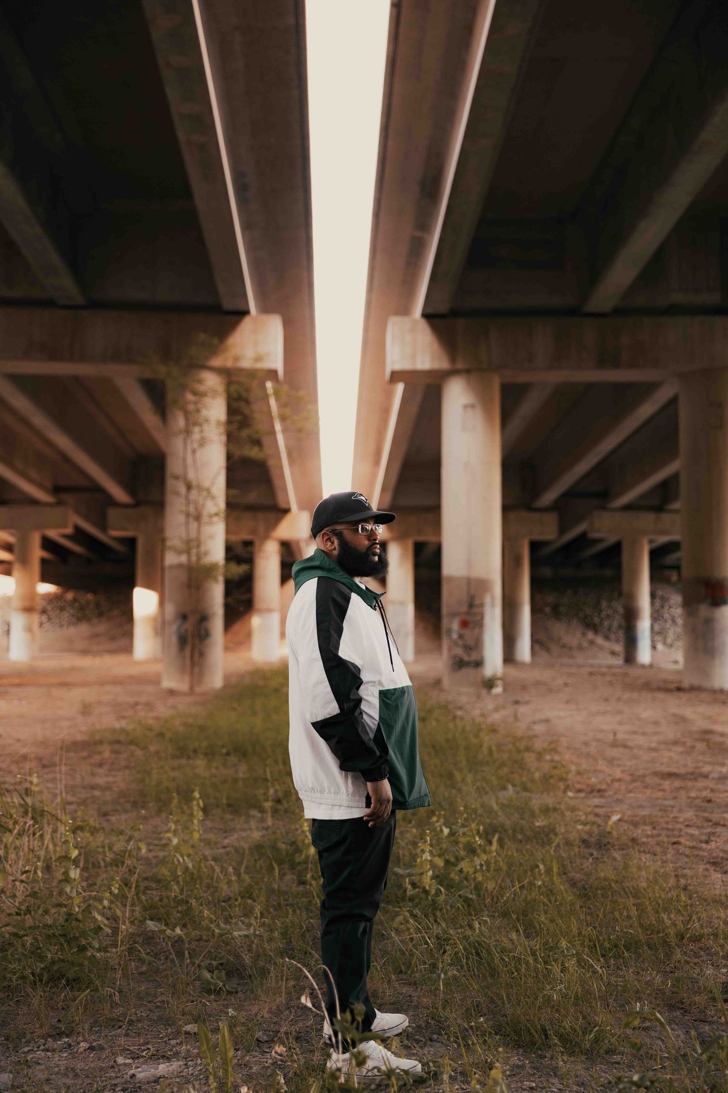 Man in streetwear standing under concrete highway overpass with graffiti columns in urban setting.
