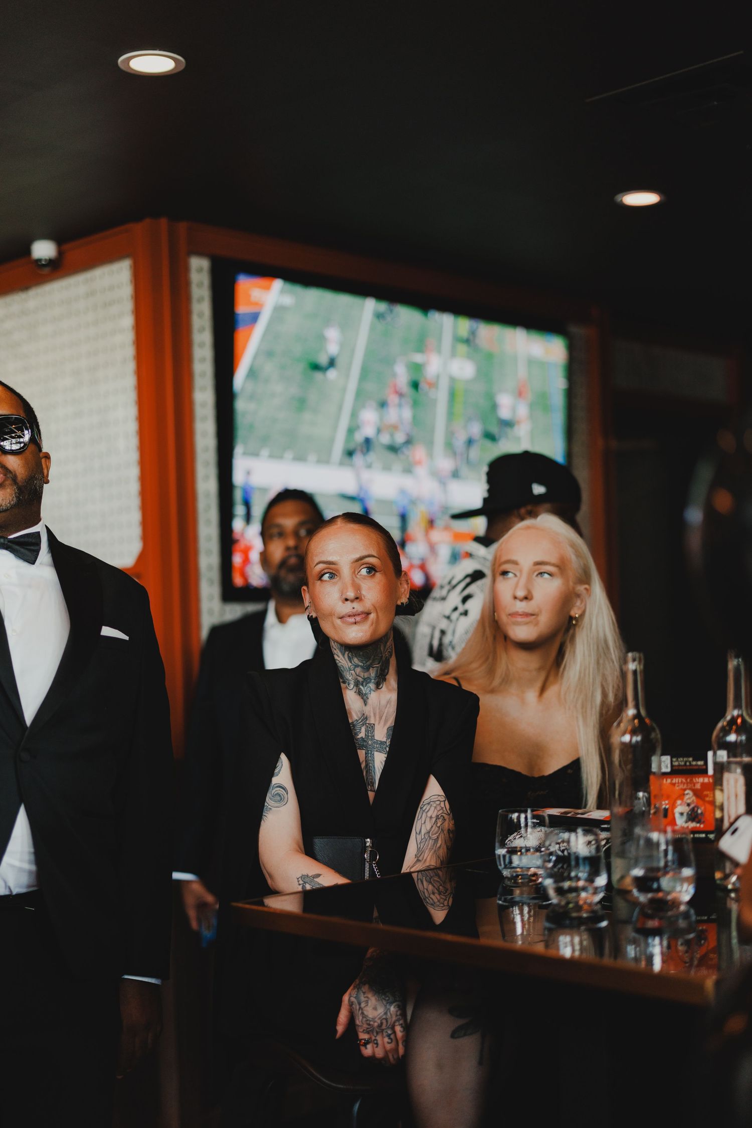 Tattooed woman in black suit with friends at a bar watching football on TV screen.