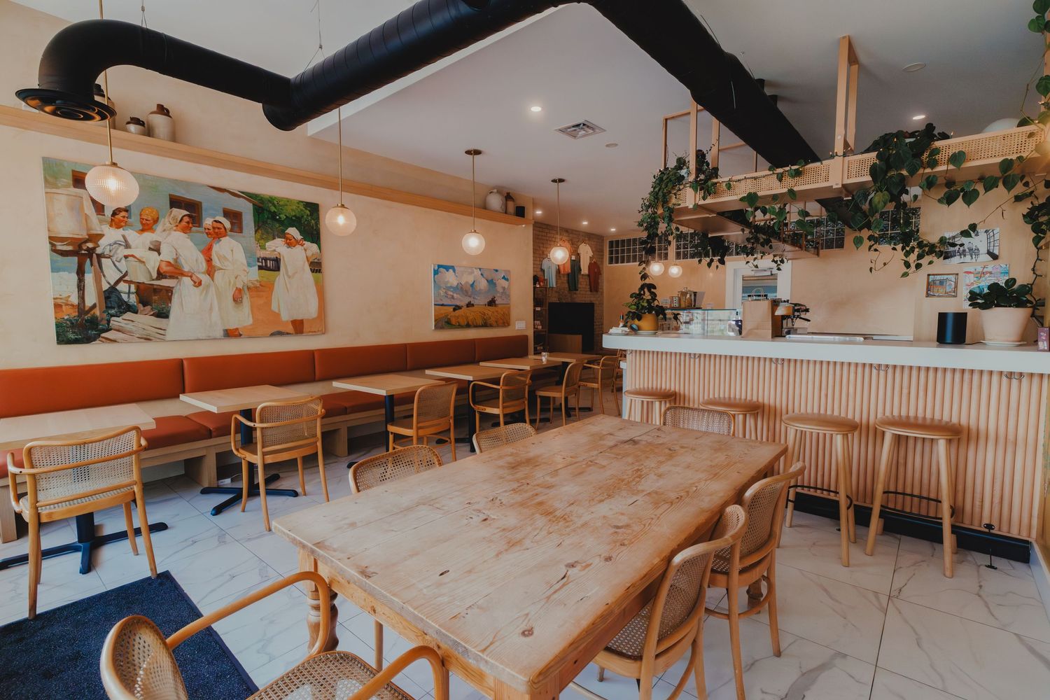 Bright modern cafe interior with wooden tables, rattan chairs, lush plants, and artwork on warm beige walls.