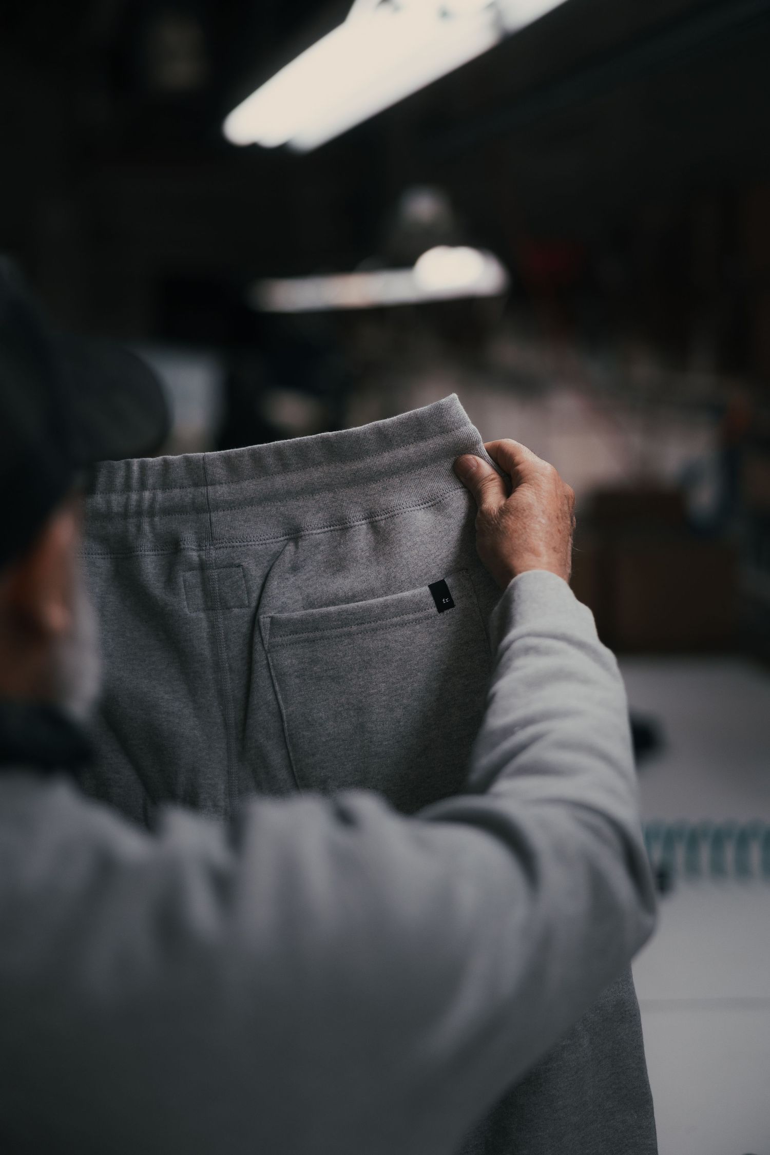 Man holding up gray fleece sweatpants with a small logo patch in an industrial workshop setting.