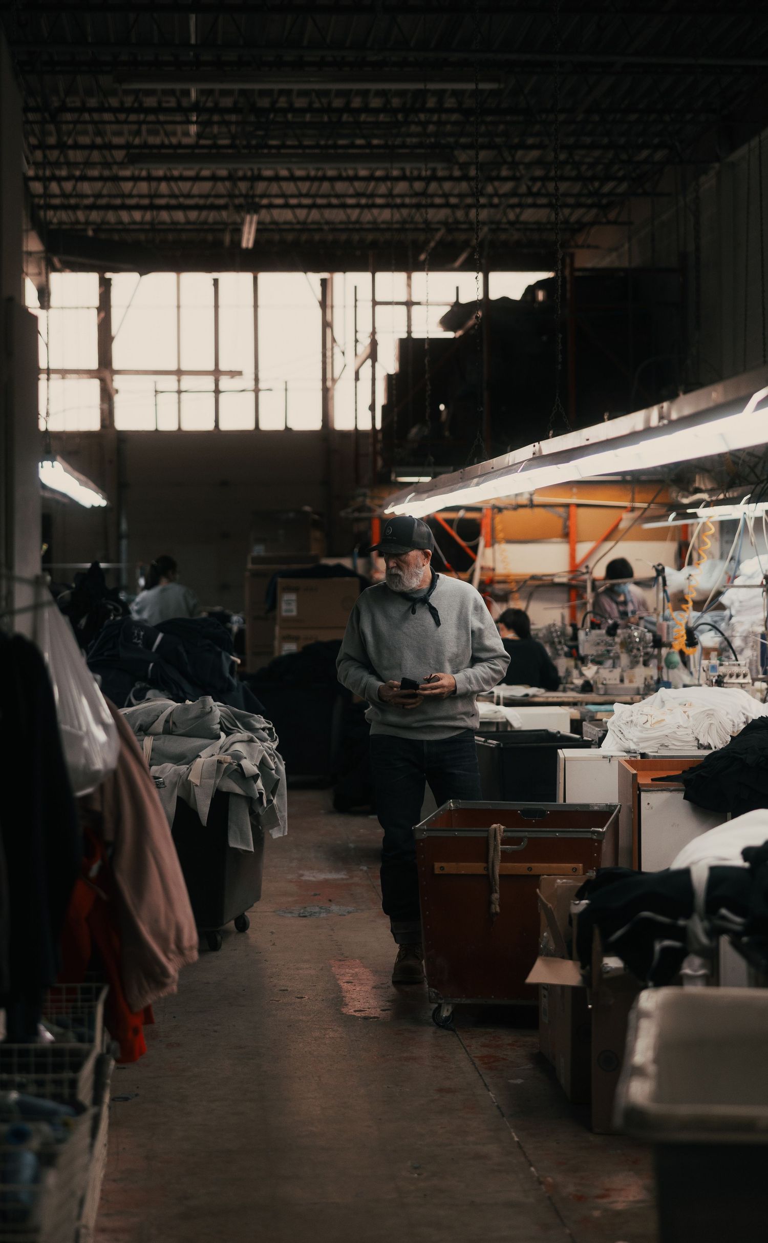Worker in a busy garment factory warehouse, surrounded by clothing racks and sewing stations under industrial lighting.
