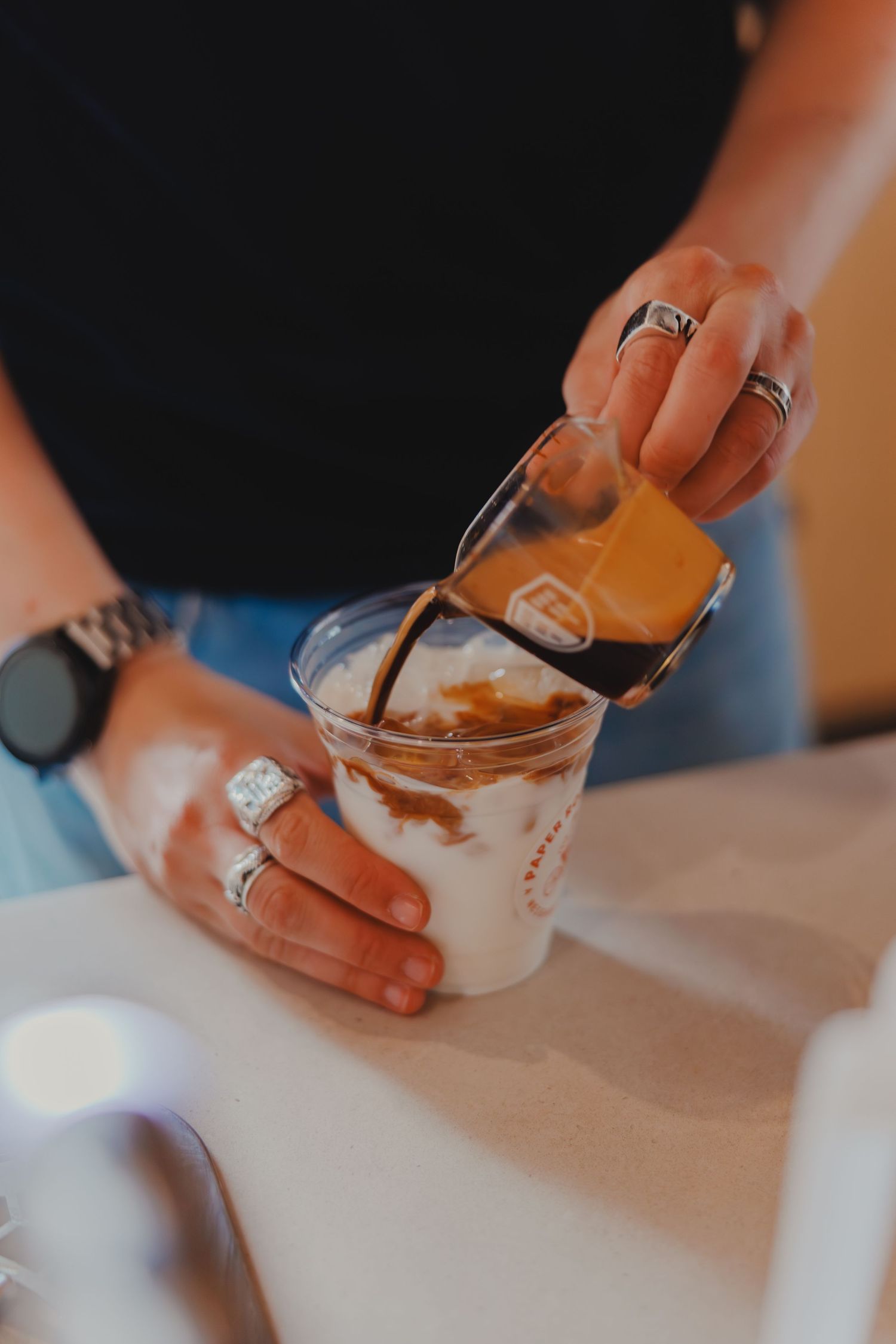Barista pouring espresso shot over ice and milk in a clear plastic cup to make an iced latte.