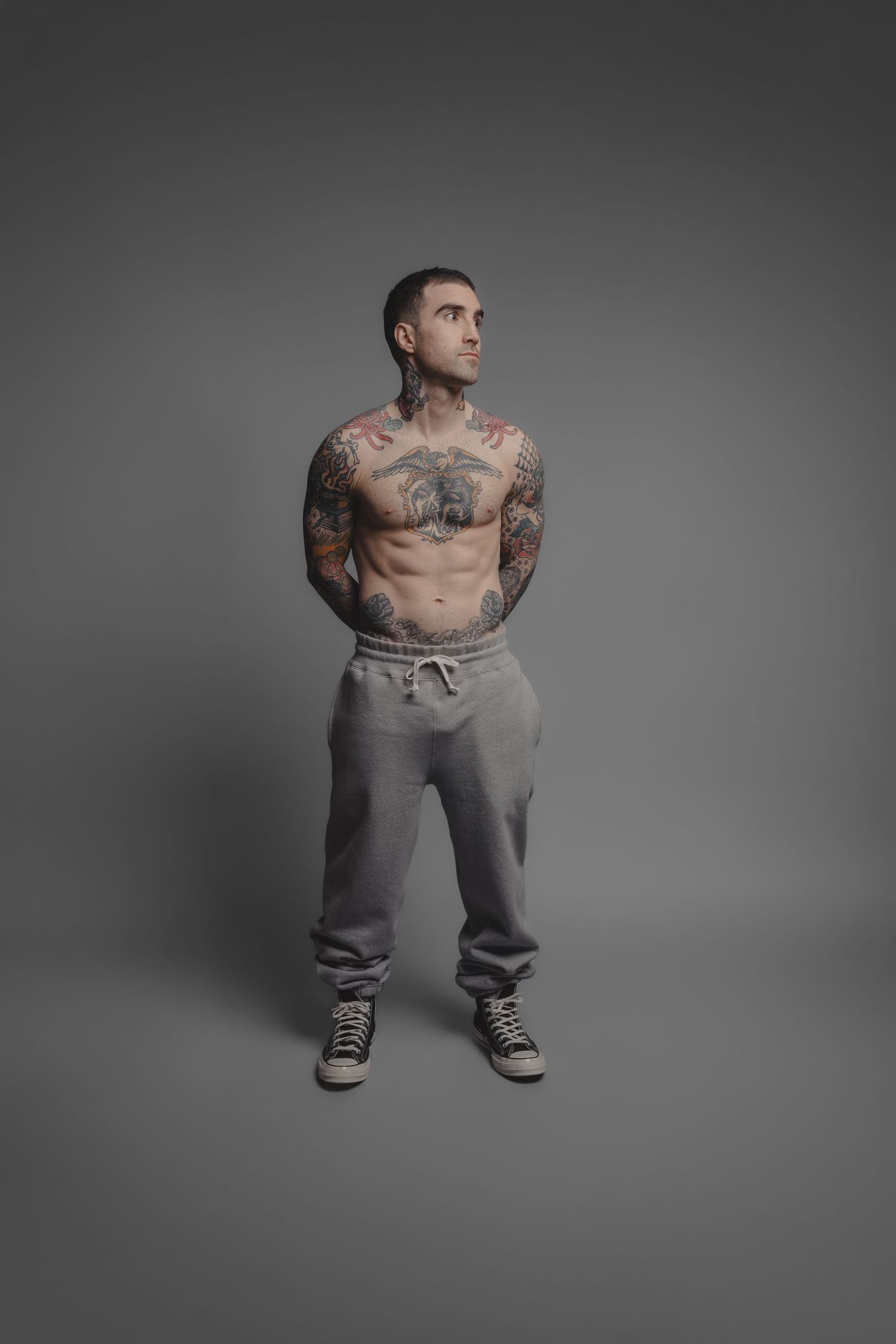 Tattooed shirtless man with toned physique wearing grey sweatpants and sneakers posing against grey background.