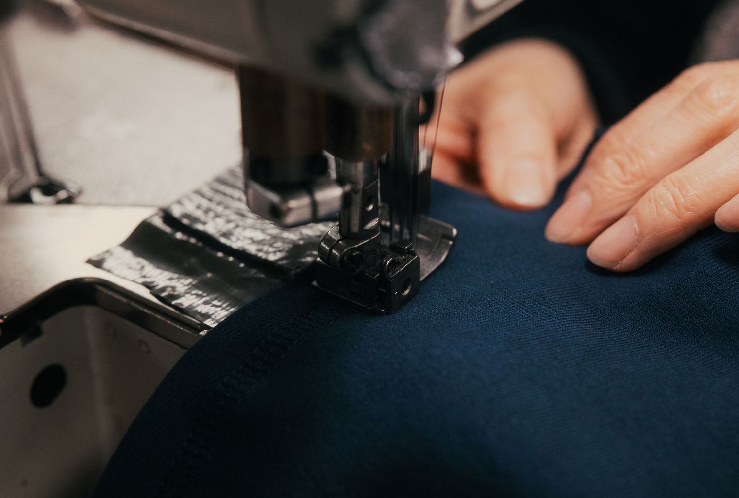 Close-up of hands guiding dark blue fabric through a sewing machine needle in a professional tailoring setting.