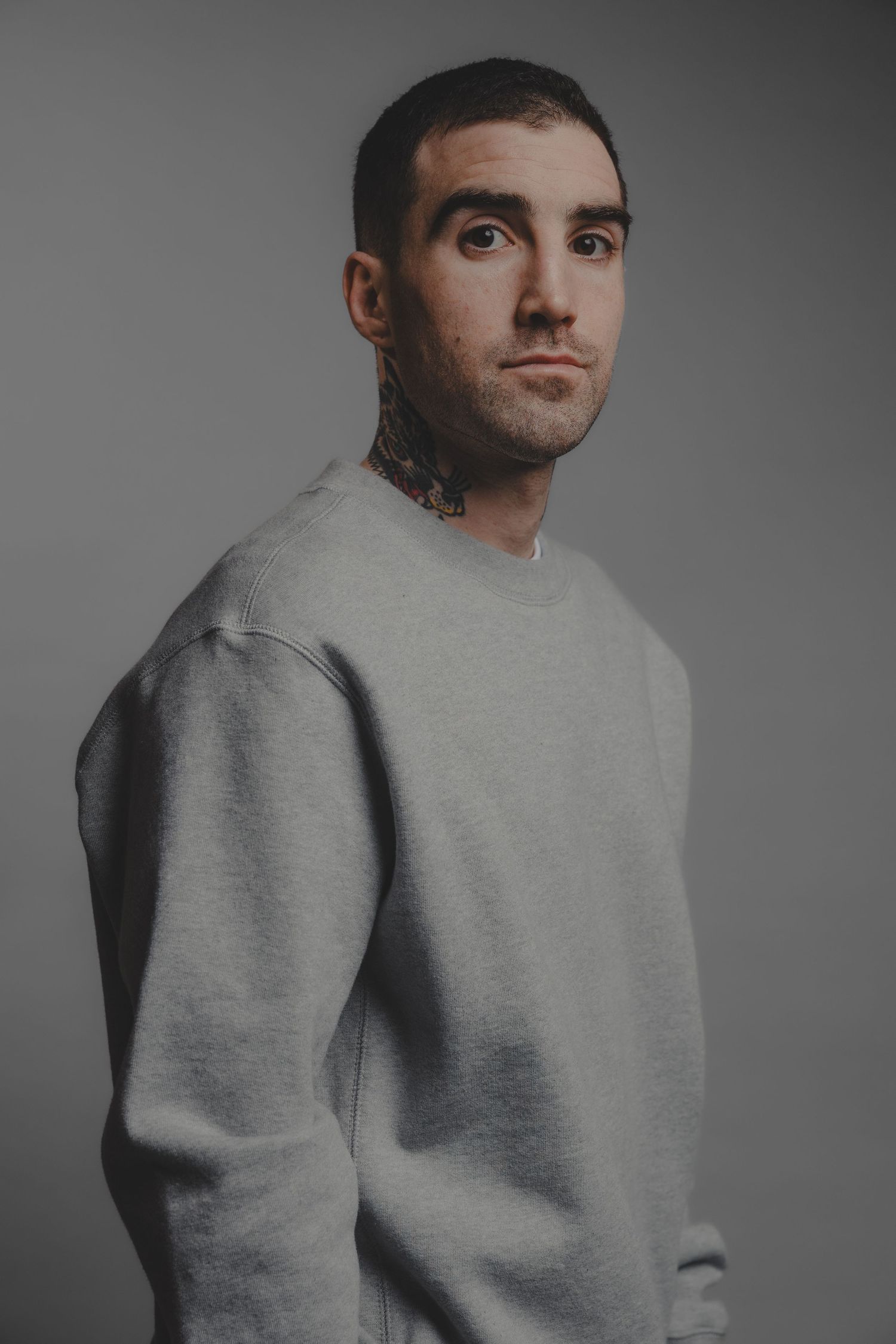 Young man with neck tattoos wearing grey crewneck sweatshirt posing against neutral grey studio background.