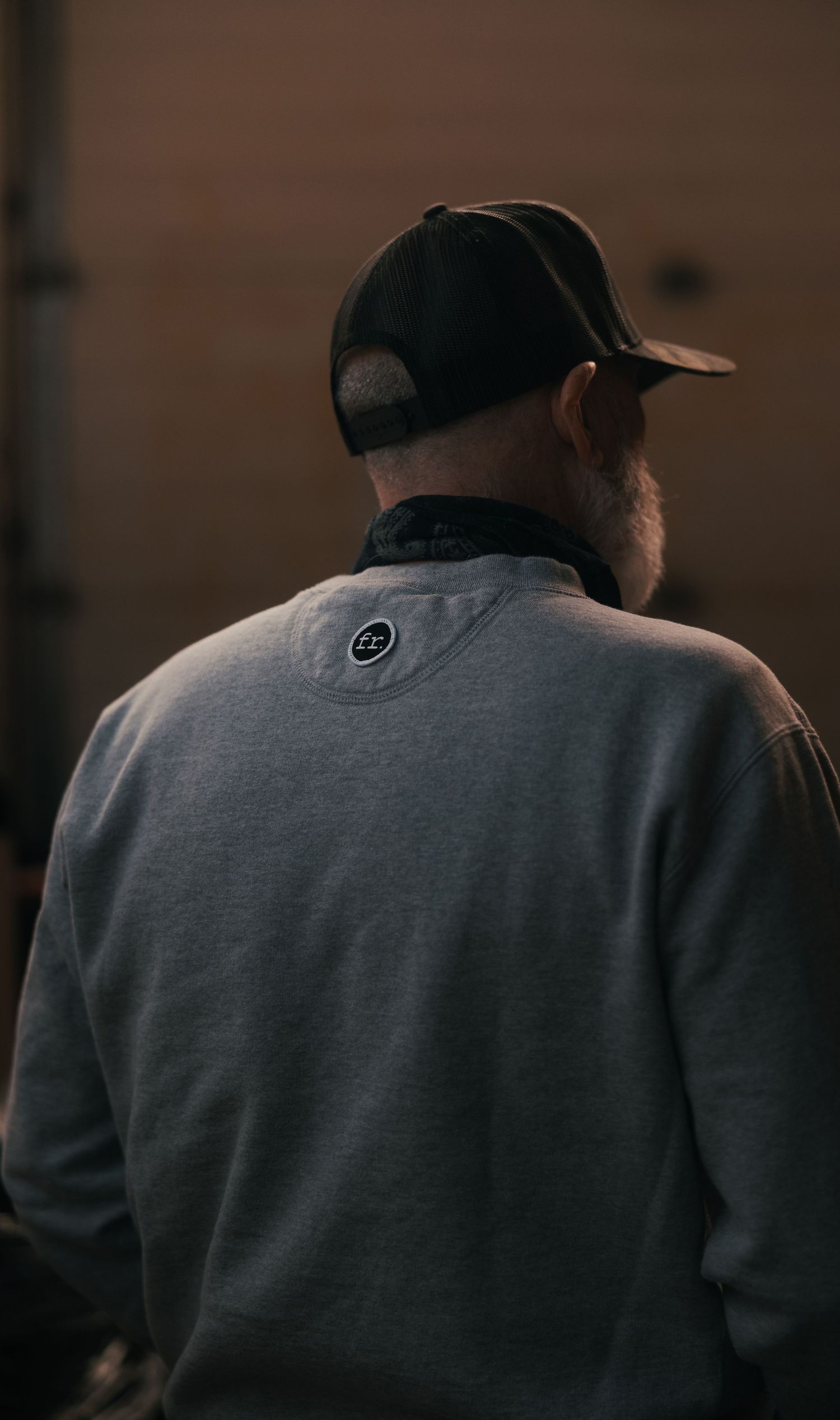 Man in gray sweatshirt and black cap seen from behind in dimly lit setting.