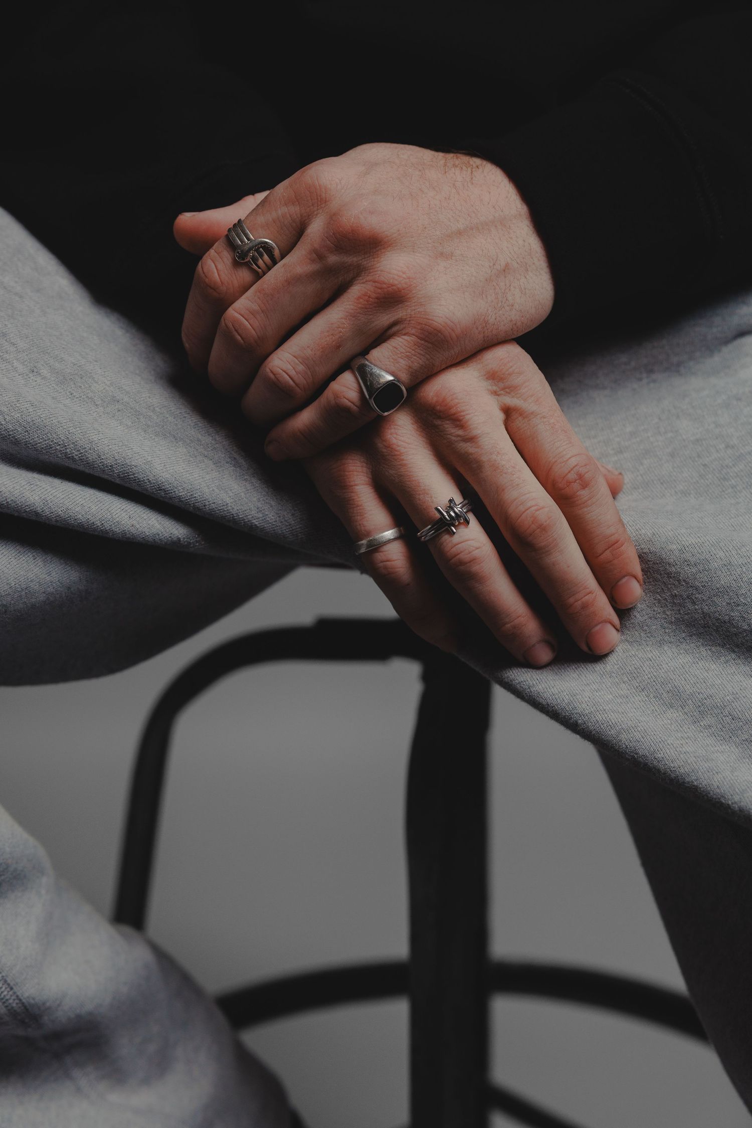 Close-up of hands wearing multiple silver rings resting on grey fabric chair in dramatic lighting.