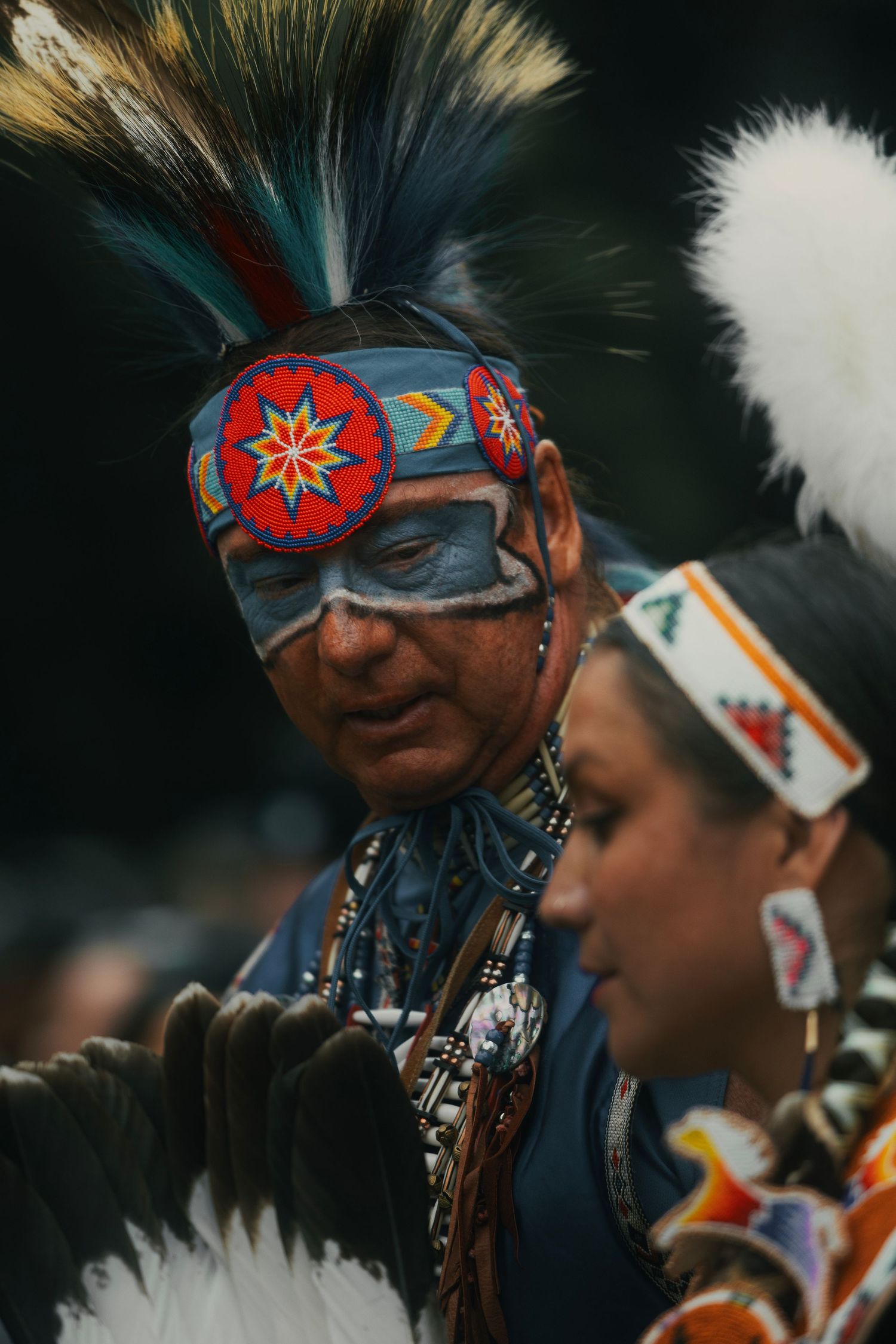 Native American man in traditional regalia with beaded headband, face paint, and feathered headdress at a powwow ceremony.