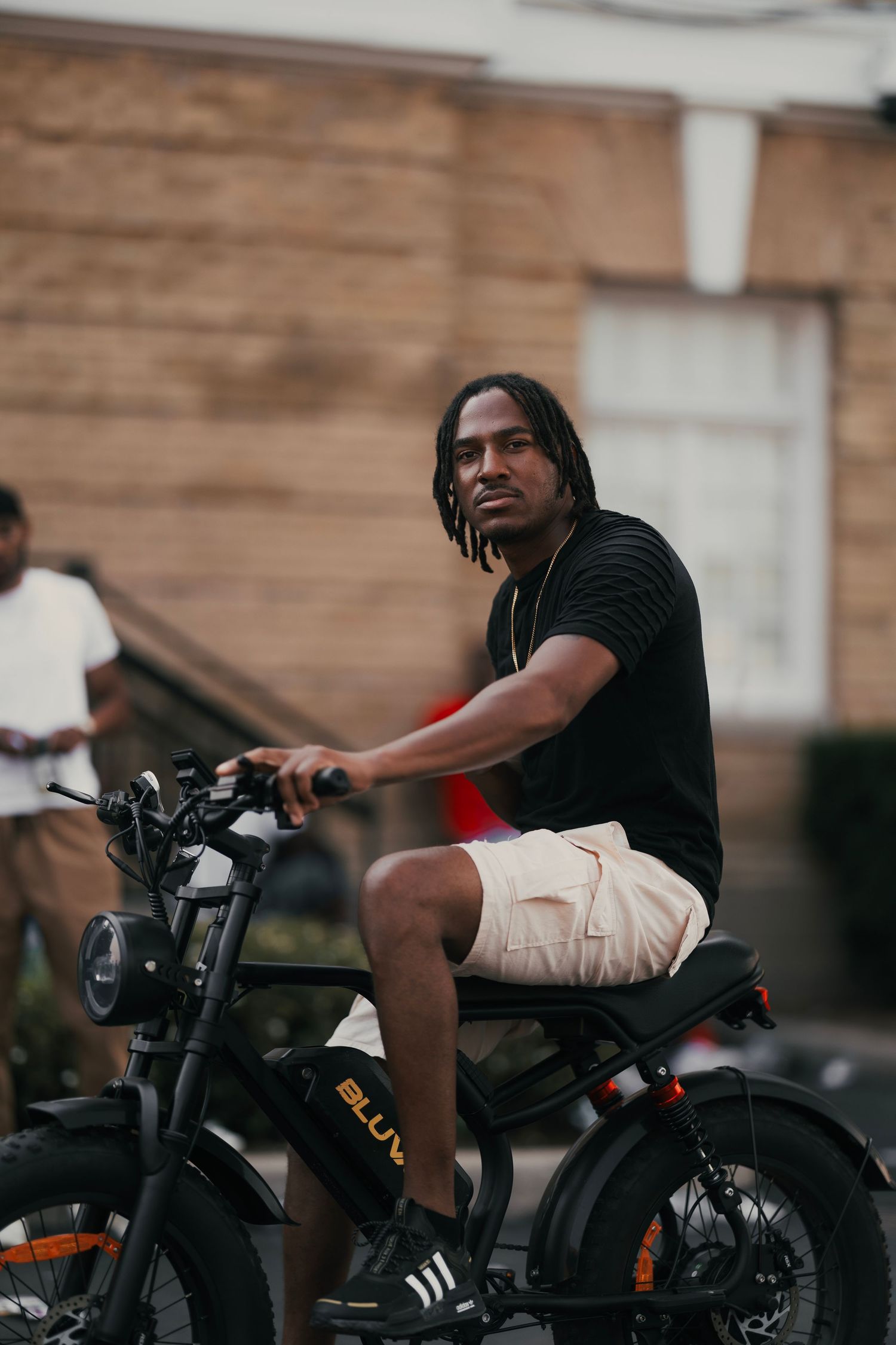 Young man with dreadlocks sitting on a black Bluuv electric bike outdoors, wearing black shirt and beige shorts.