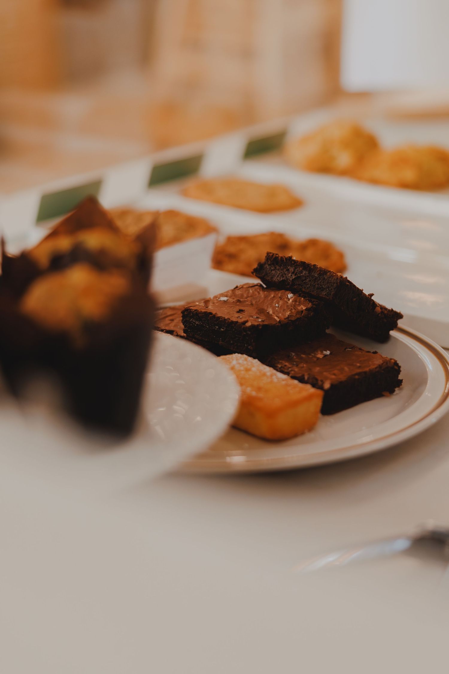 A white plate with chocolate brownies and golden baked goods displayed on a white table in a cafe.