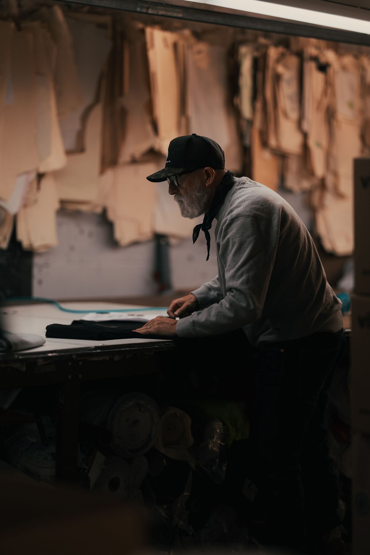 Elderly craftsman in black cap and glasses works at a leather workshop bench surrounded by hanging hides.