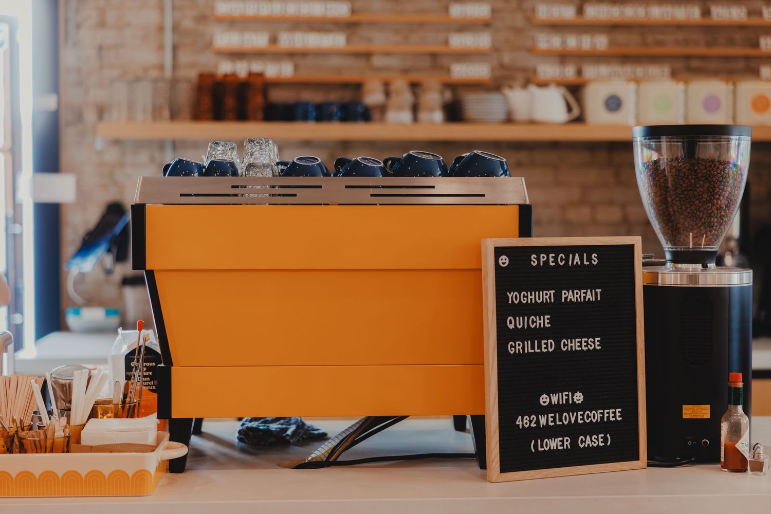 Bright orange espresso machine on cafe counter with specials chalkboard sign and coffee grinder.