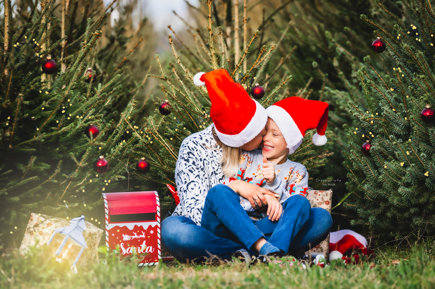 Couple in Santa hats sit together among Christmas trees surrounded by colorful wrapped presents.