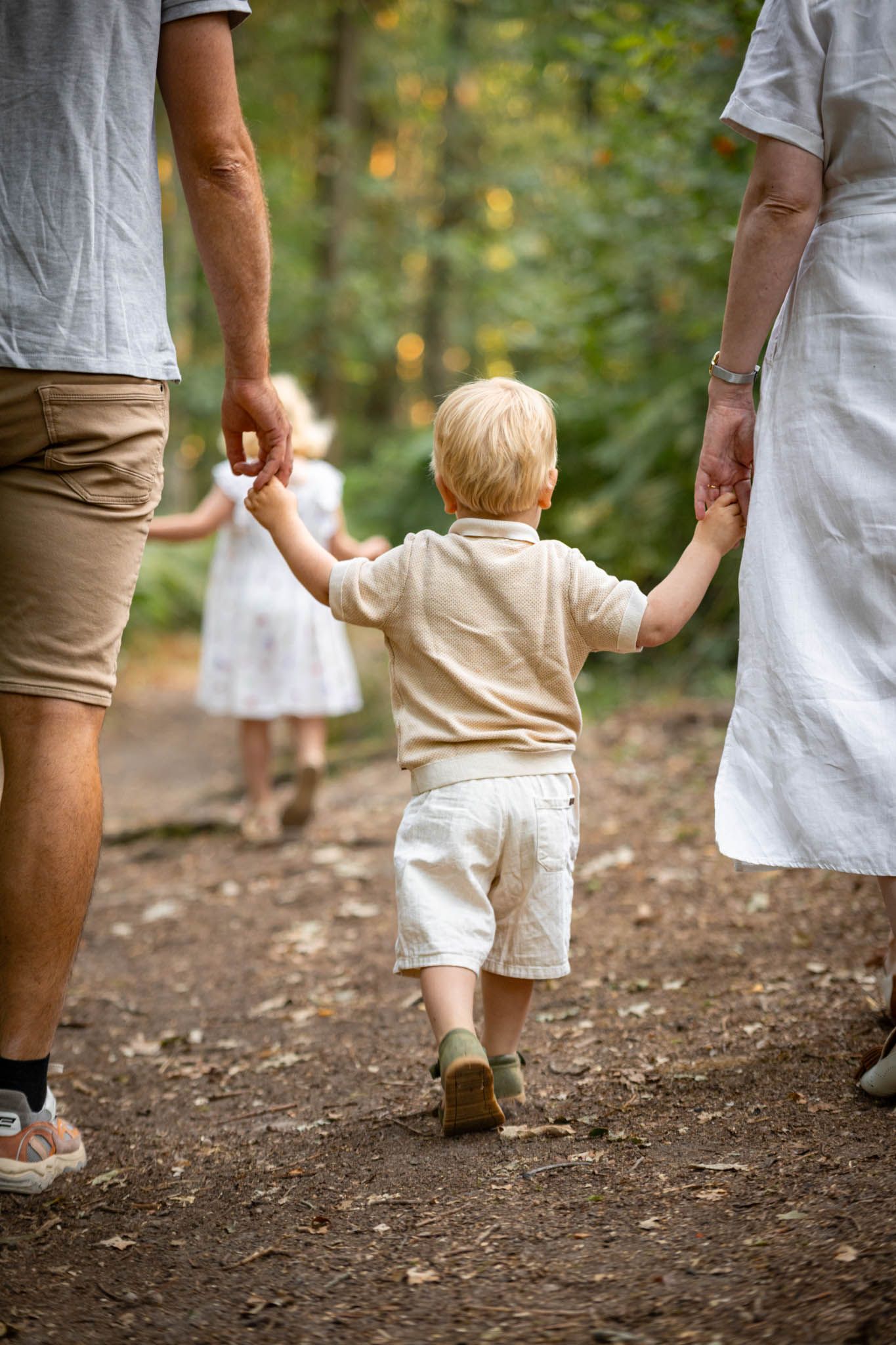 A young toddler boy holding hands with parents while walking on a forest path with a girl ahead.