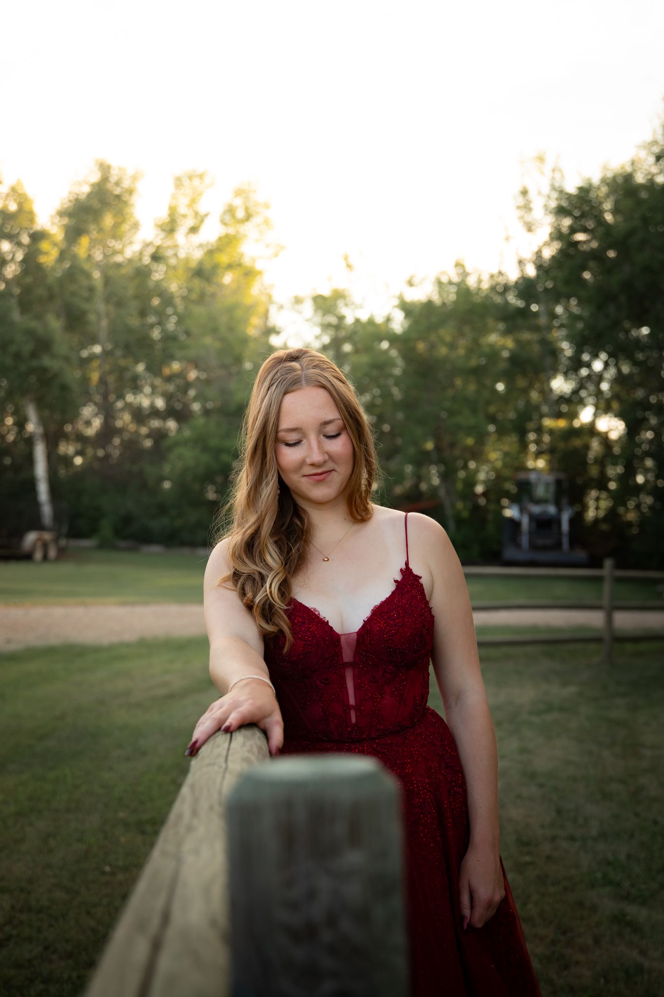 A person in a burgundy dress poses in front of a grassy outdoor setting at sunset.