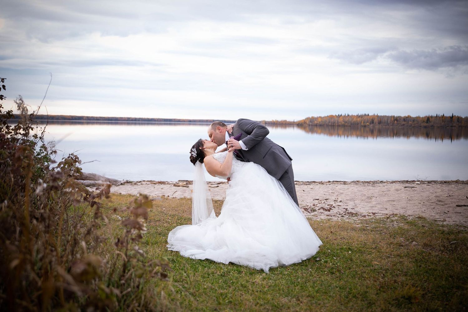 A wedding couple poses near a scenic lake shore on an overcast day.