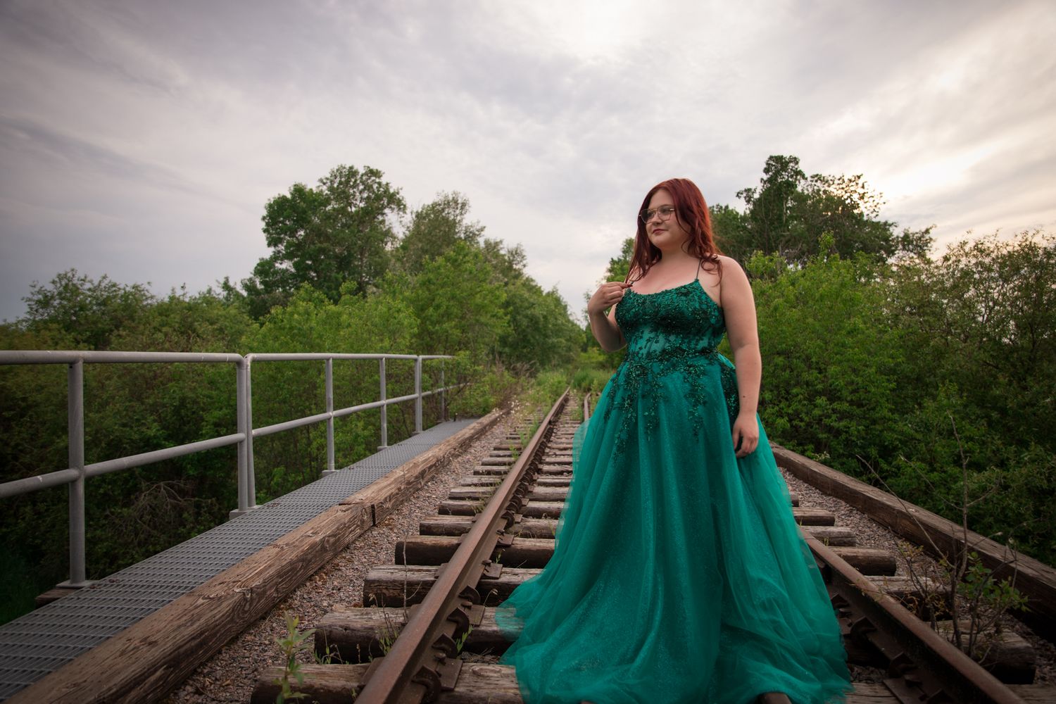 Elegant emerald ball gown photographed on rustic railroad bridge at sunset.