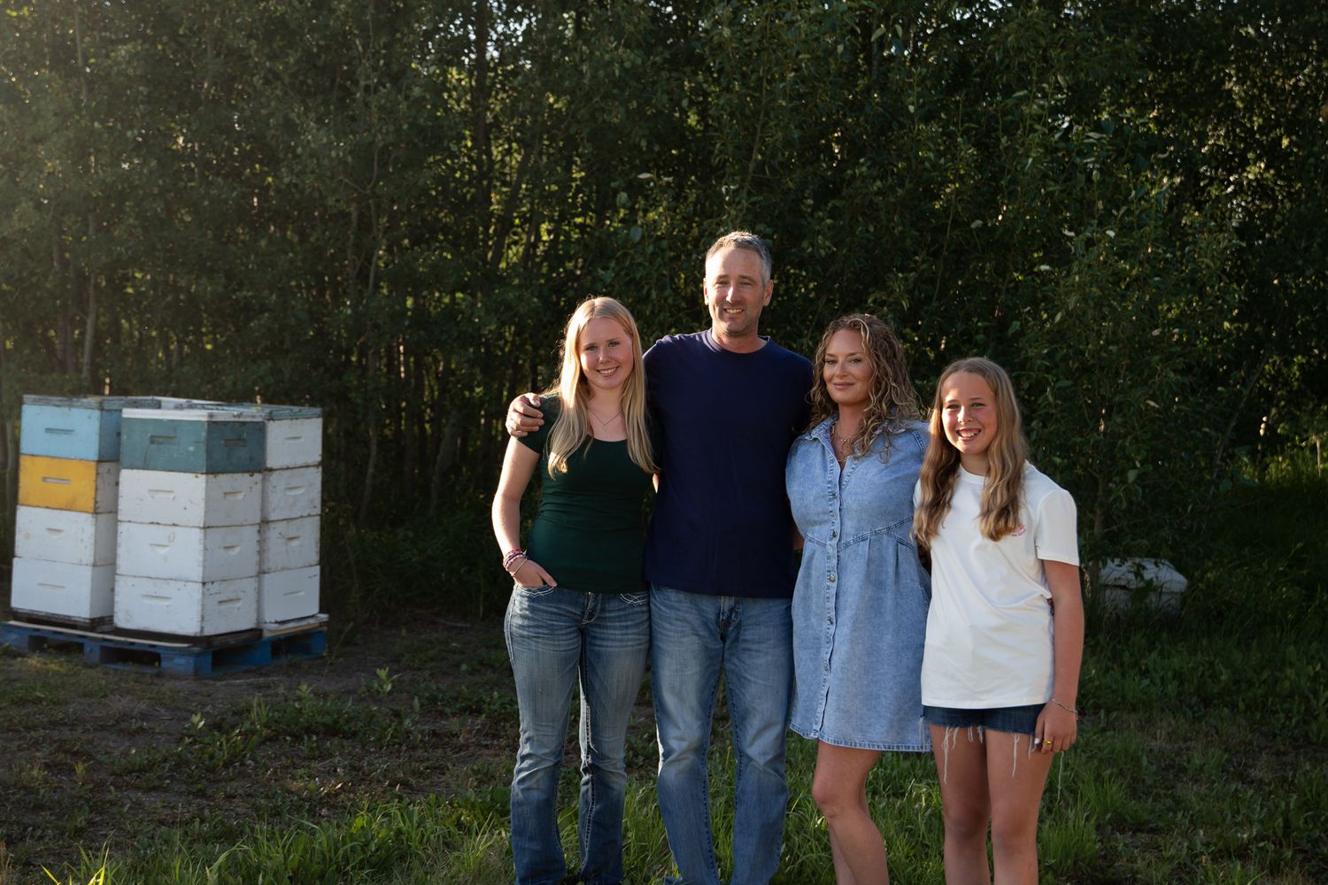 A group of people stand near beehive boxes at the edge of a wooded area.