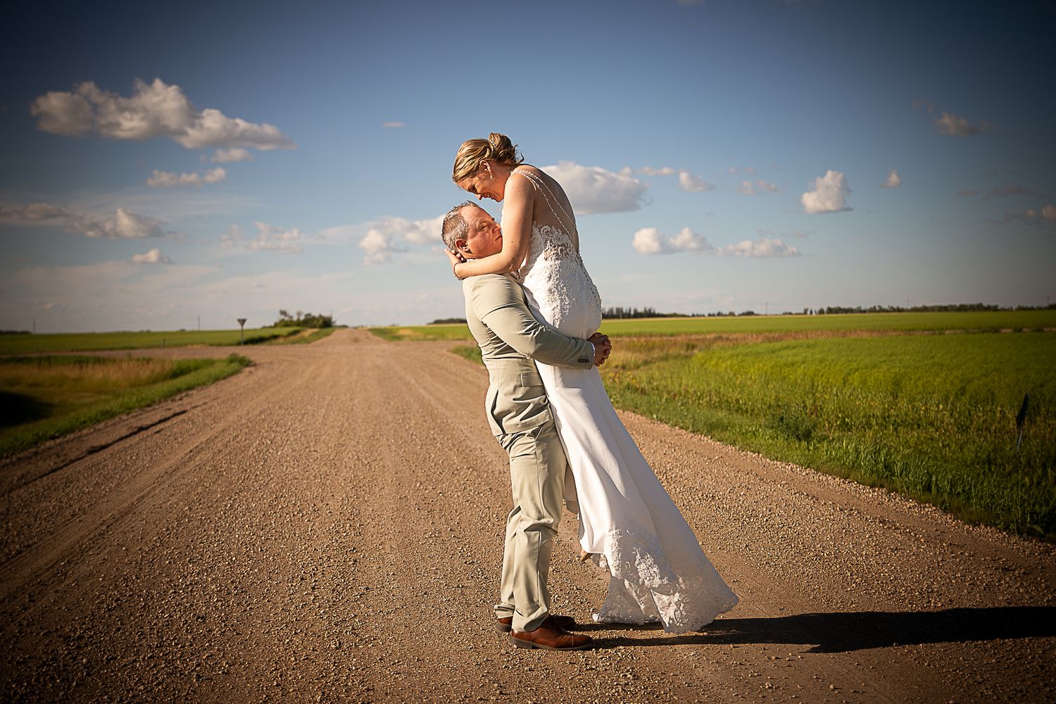 Wedding couple shares romantic moments on rural dirt road surrounded by farmland.