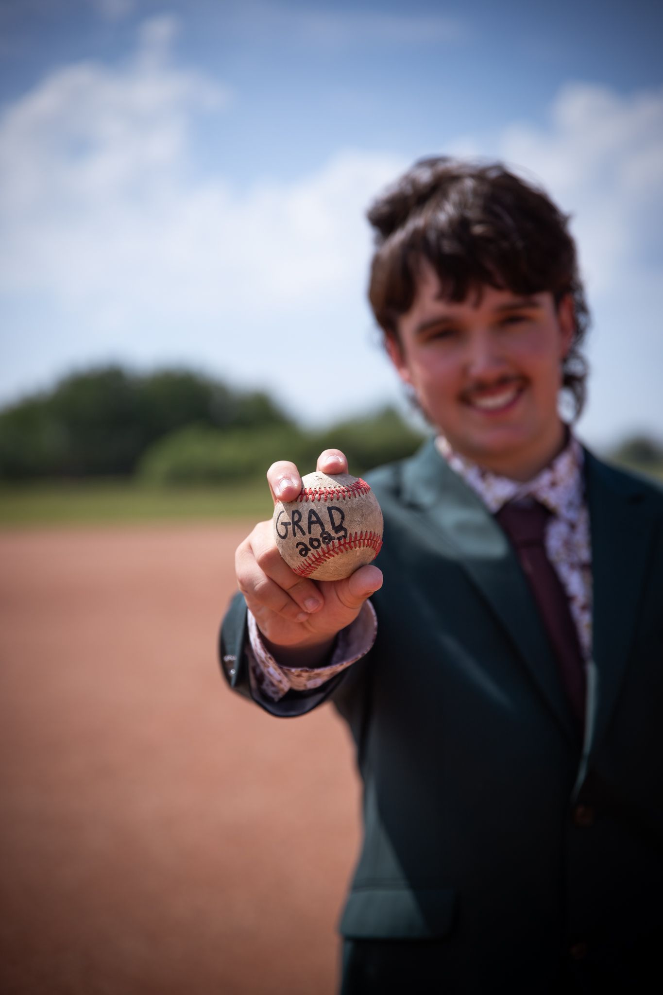 A hand holds up a baseball while wearing a formal dark green suit.