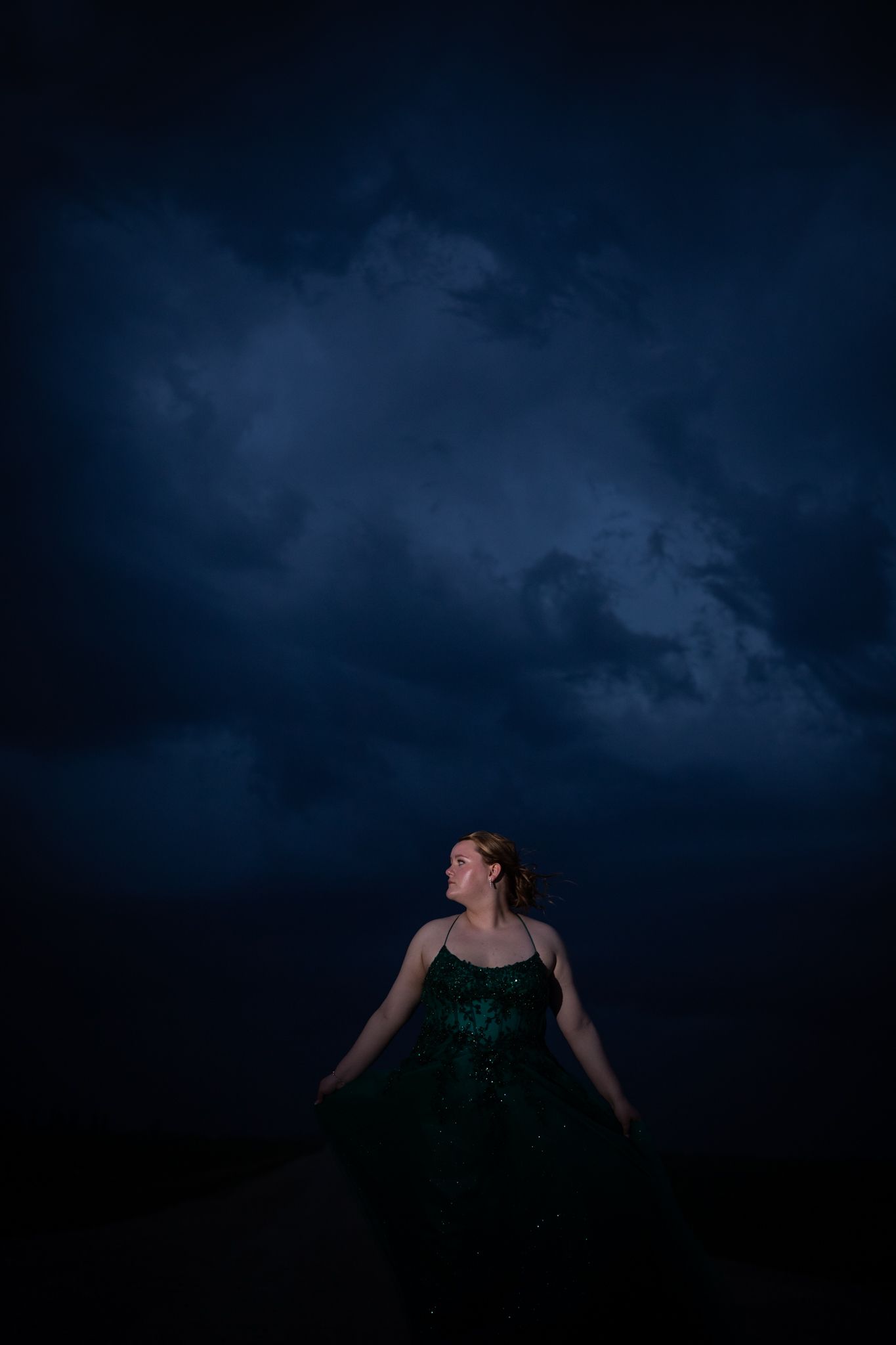 A dramatic pose in an emerald gown is captured against dark storm clouds.