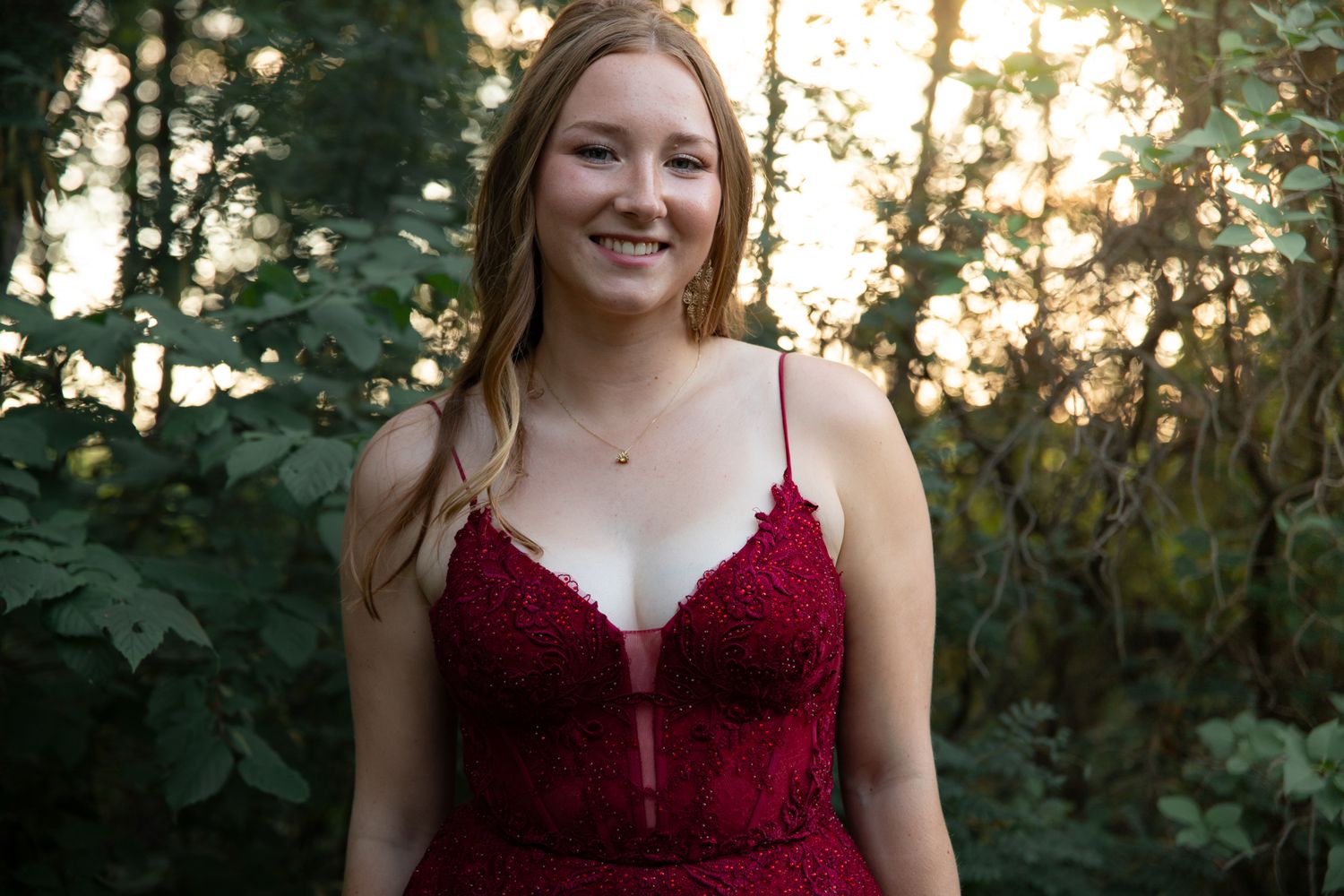 A deep red formal dress photographed in natural evening light among trees.