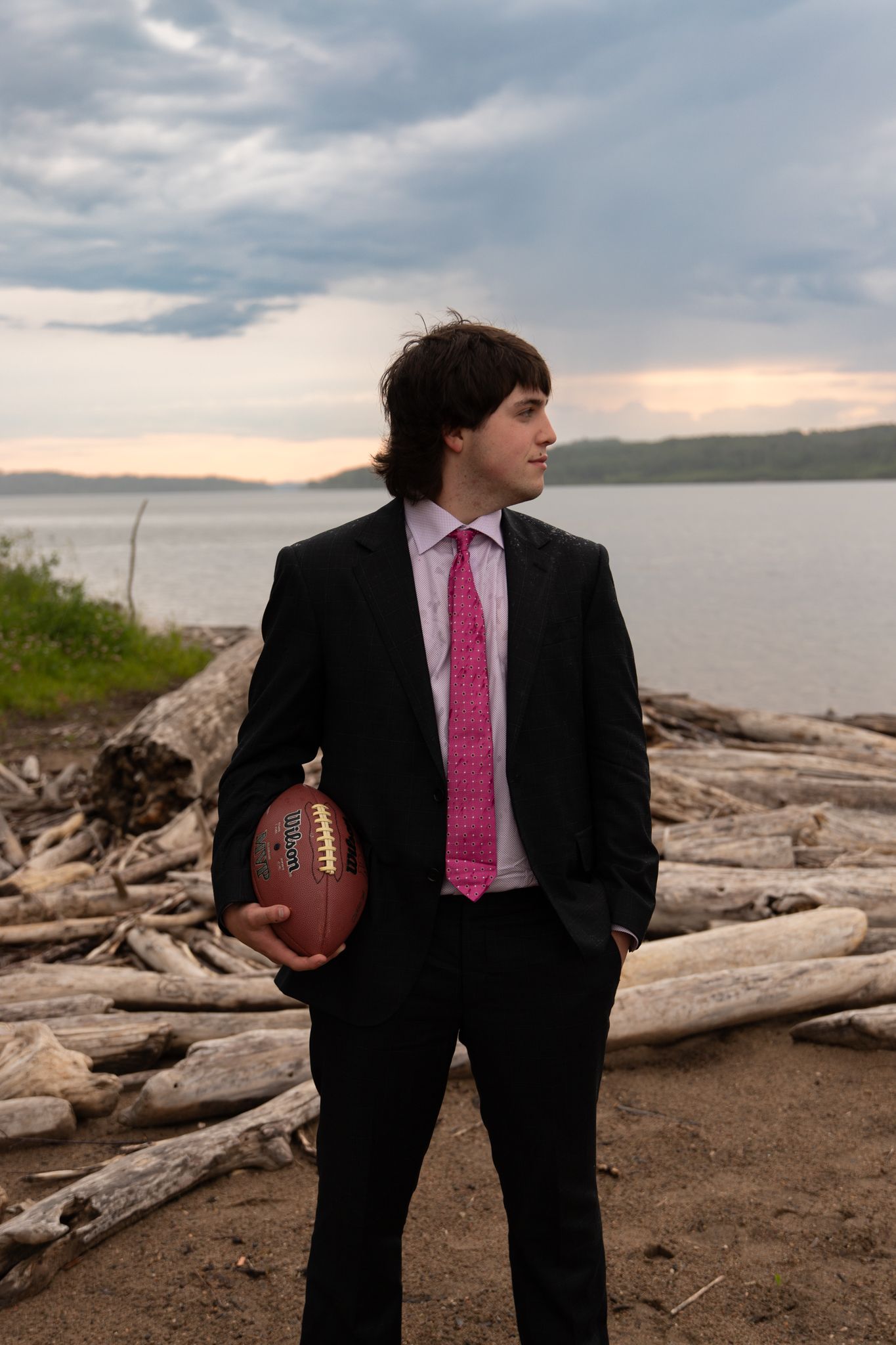A figure in a suit holds a football while standing among driftwood on a rocky shoreline.