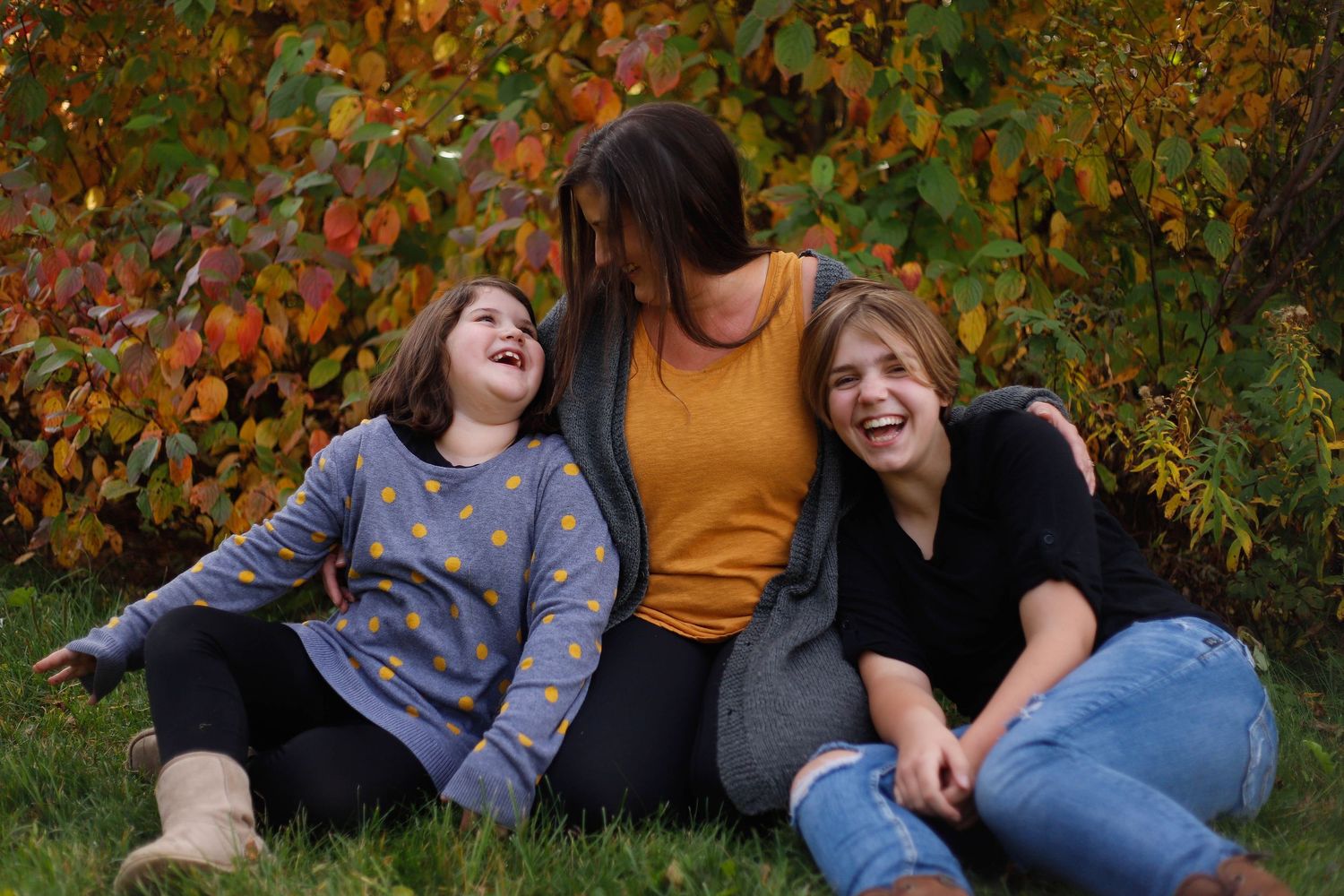 Three people laughing together while sitting on grass with colorful fall leaves in the background.