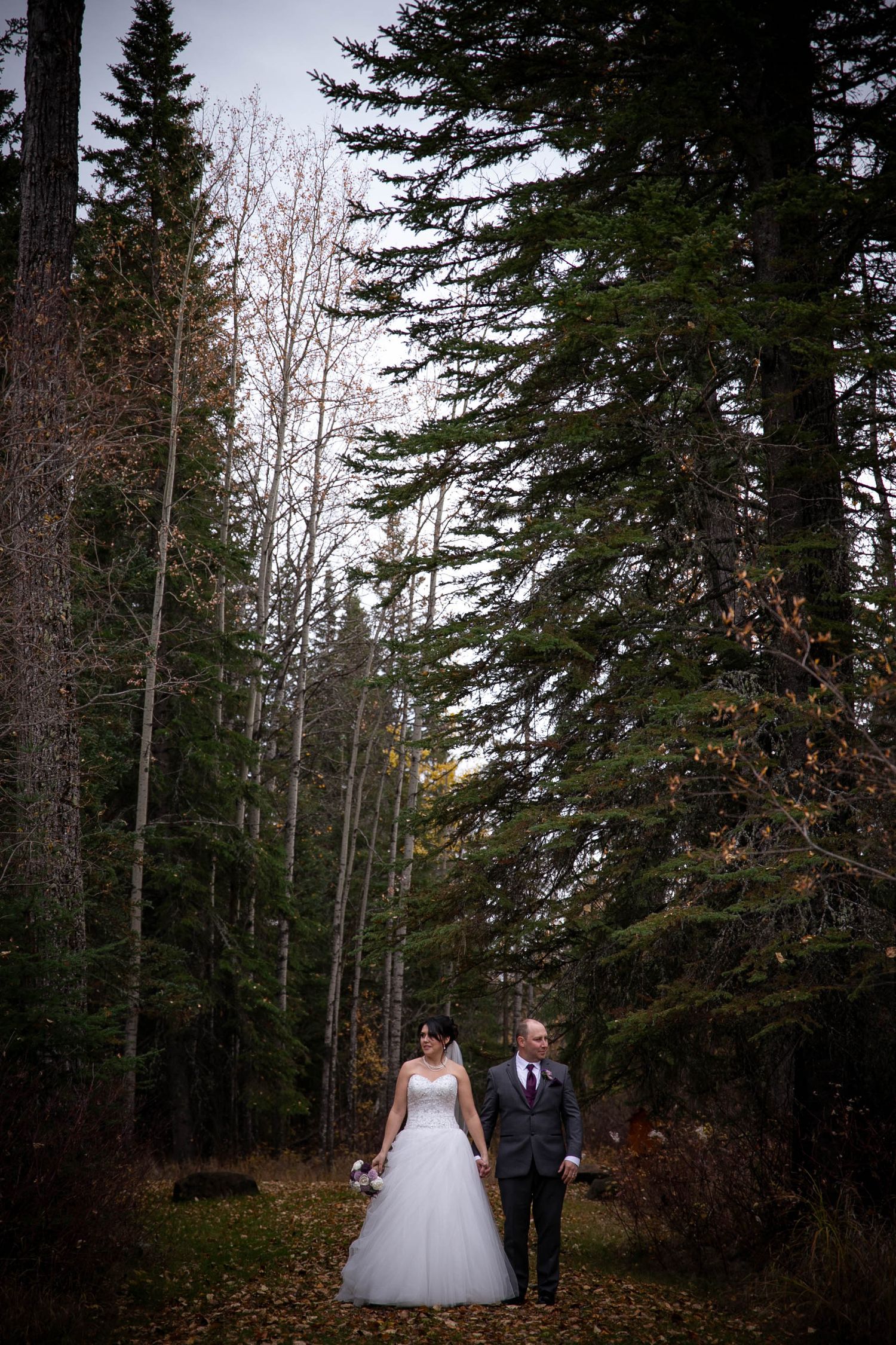 A couple walks hand in hand through a forest path in winter wedding attire.